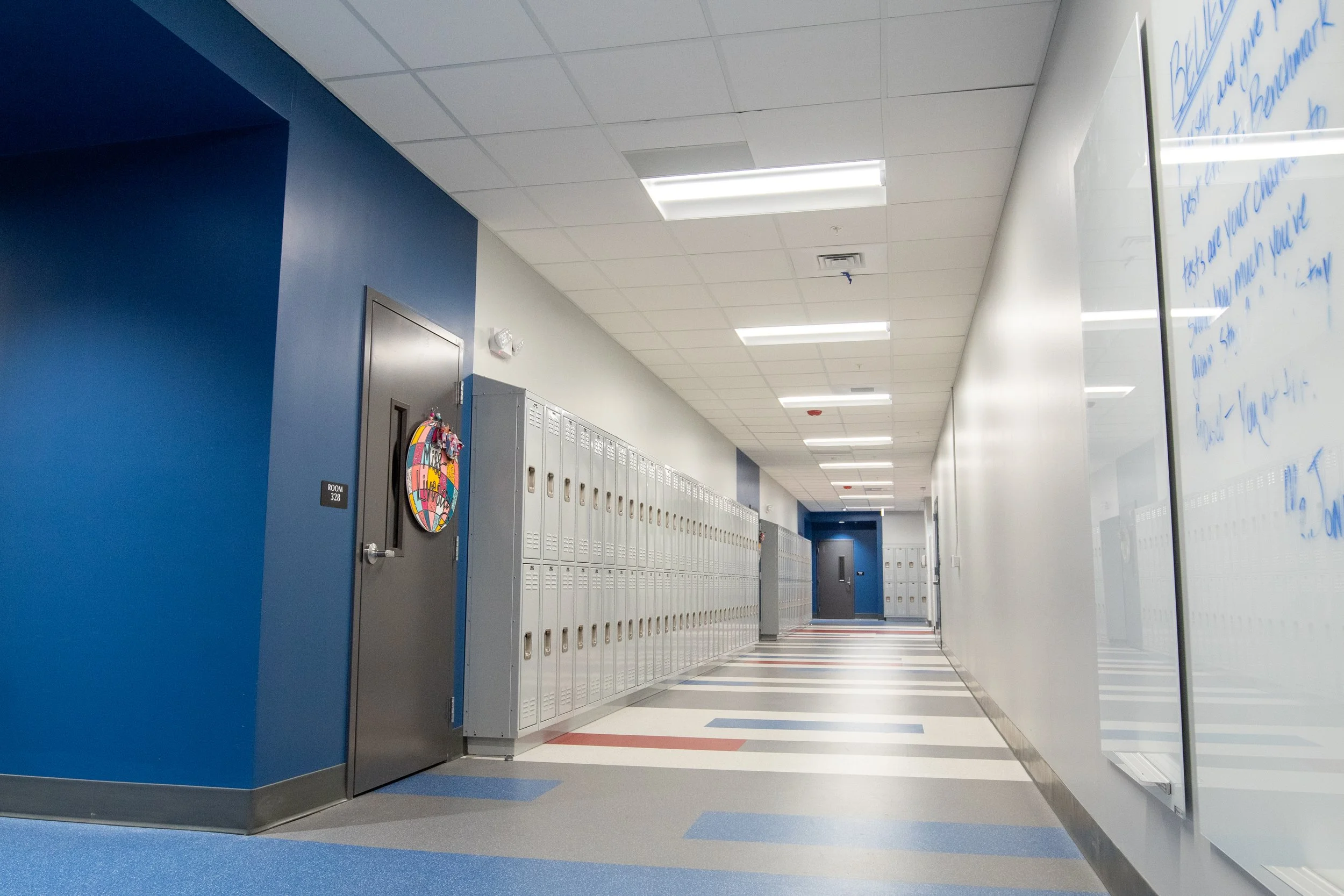Empty school hallway with blue and white walls, lockers along the side, and a whiteboard with writing on the right wall.