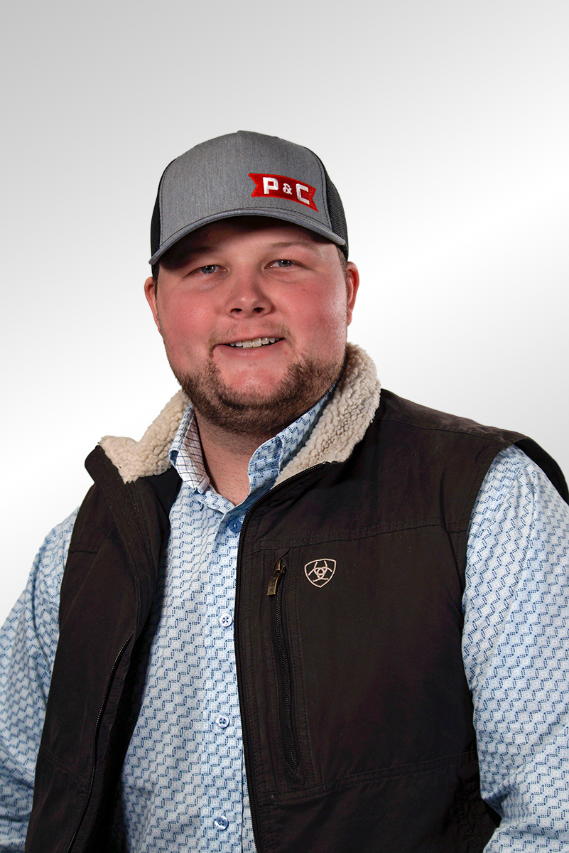 A man smiling, wearing a gray baseball cap with a red and white logo, a black vest with a zippered pocket, and a light blue patterned button-up shirt, standing against a plain white background.