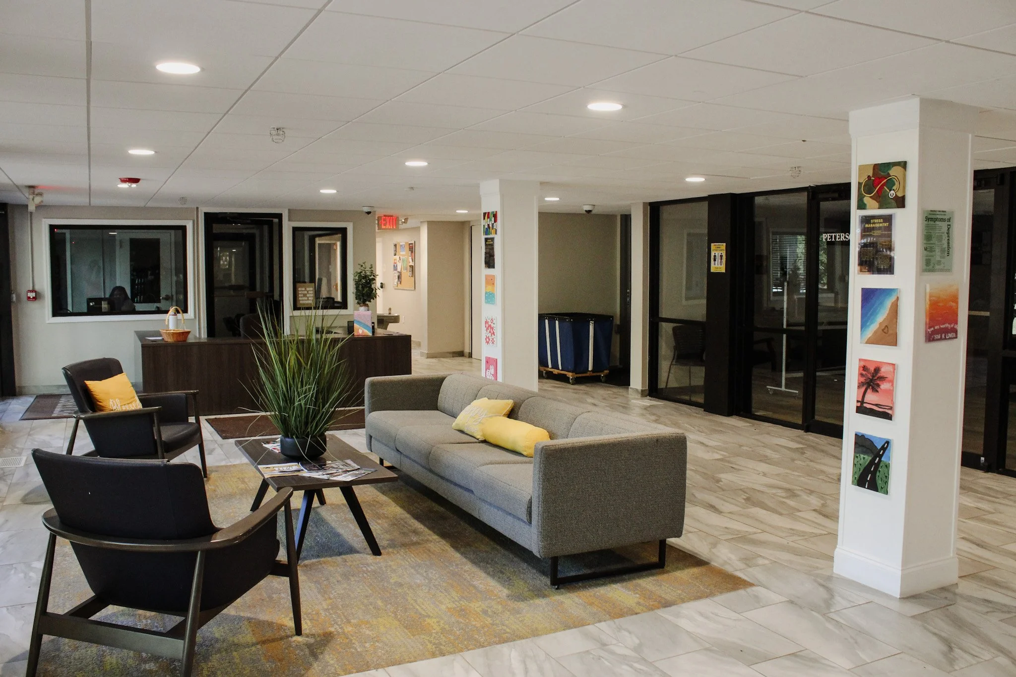 A spacious lobby area with comfortable seating including gray sofa and black chairs with yellow cushions. There are potted plants, artwork on the walls, and large windows with glass doors leading outside. The ceiling has recessed lighting and the flo