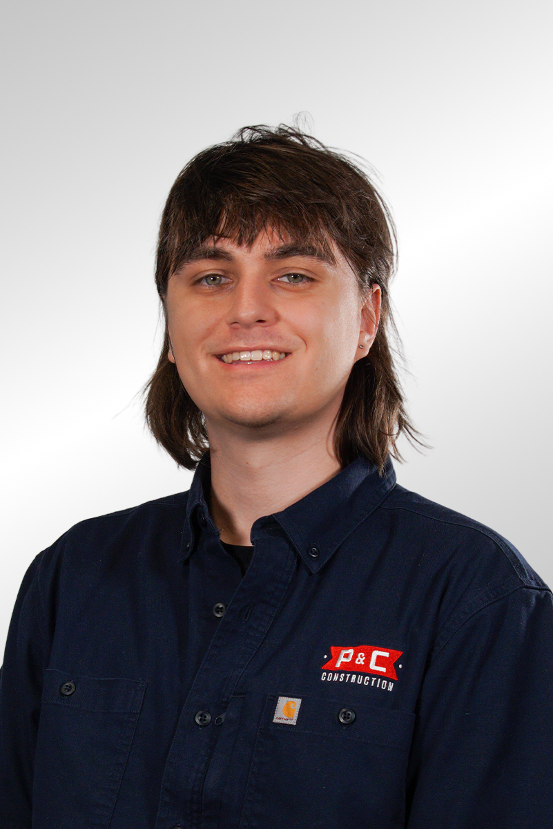 A young woman with long brown hair, smiling, wearing a pink t-shirt and a delicate necklace, standing against a dark background.