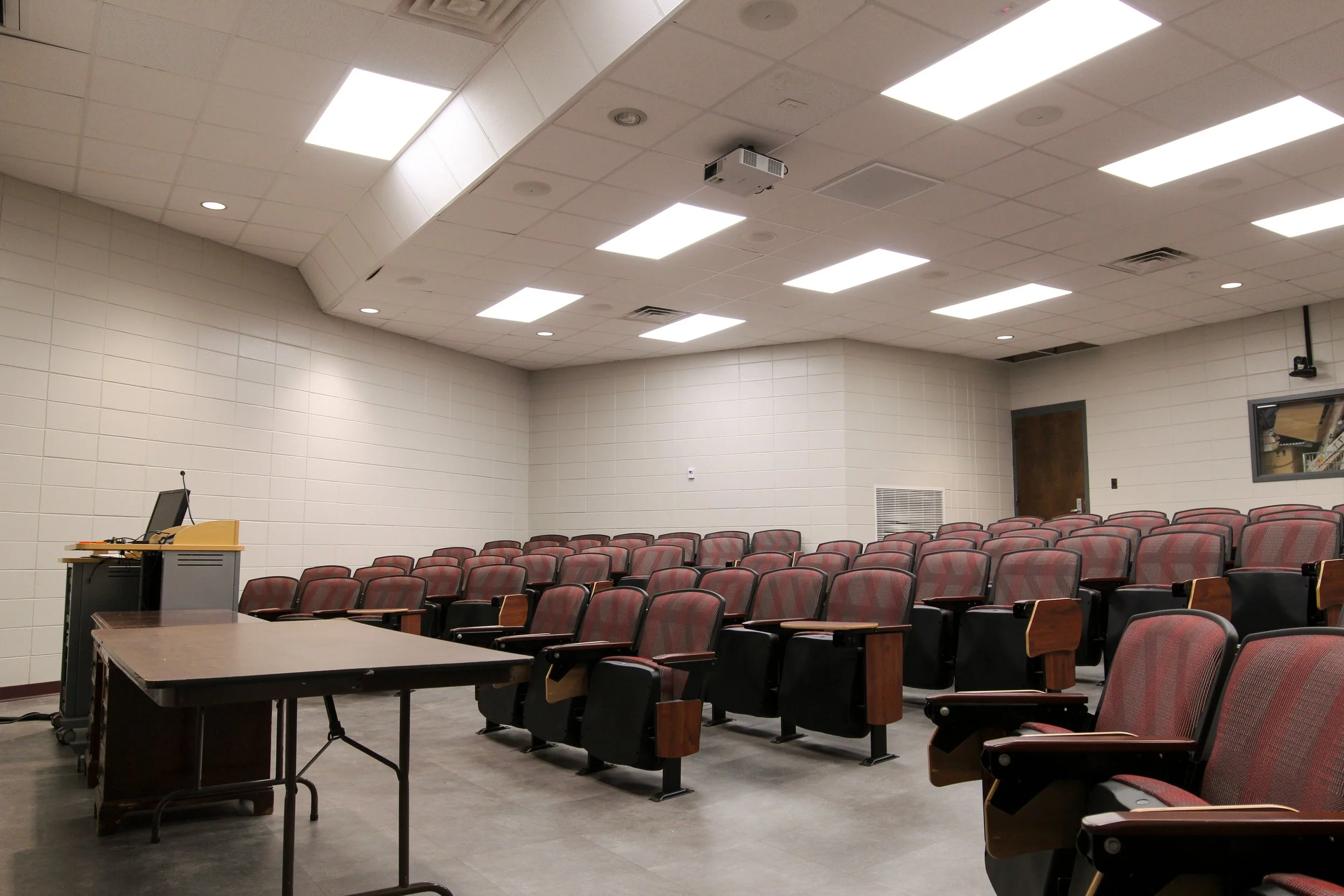 Empty lecture hall with rows of red and black seats, a wooden desk at the front, and white tiled walls and ceiling.