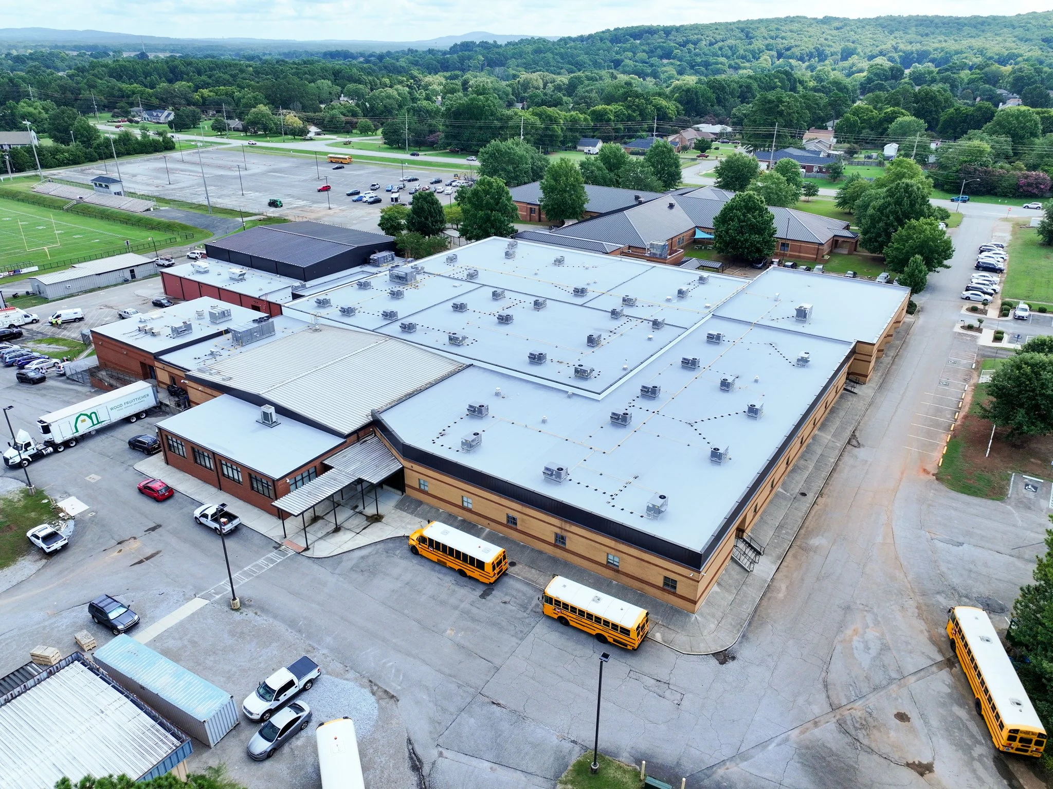 Aerial view of Discovery Middle School with a flat roof equipped with HVAC units, adjacent parking lot with cars, surrounding greenery, and a distant landscape of trees and hills.