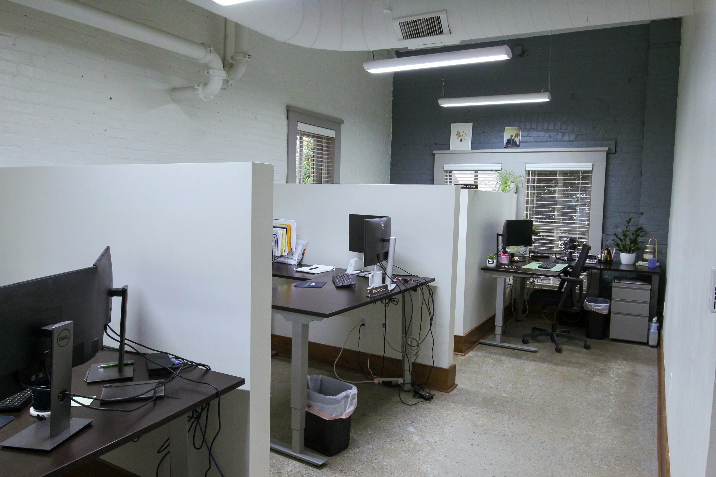 Office space with desks, computers, and plants, separated by white partitions, with a dark brick wall and large windows.