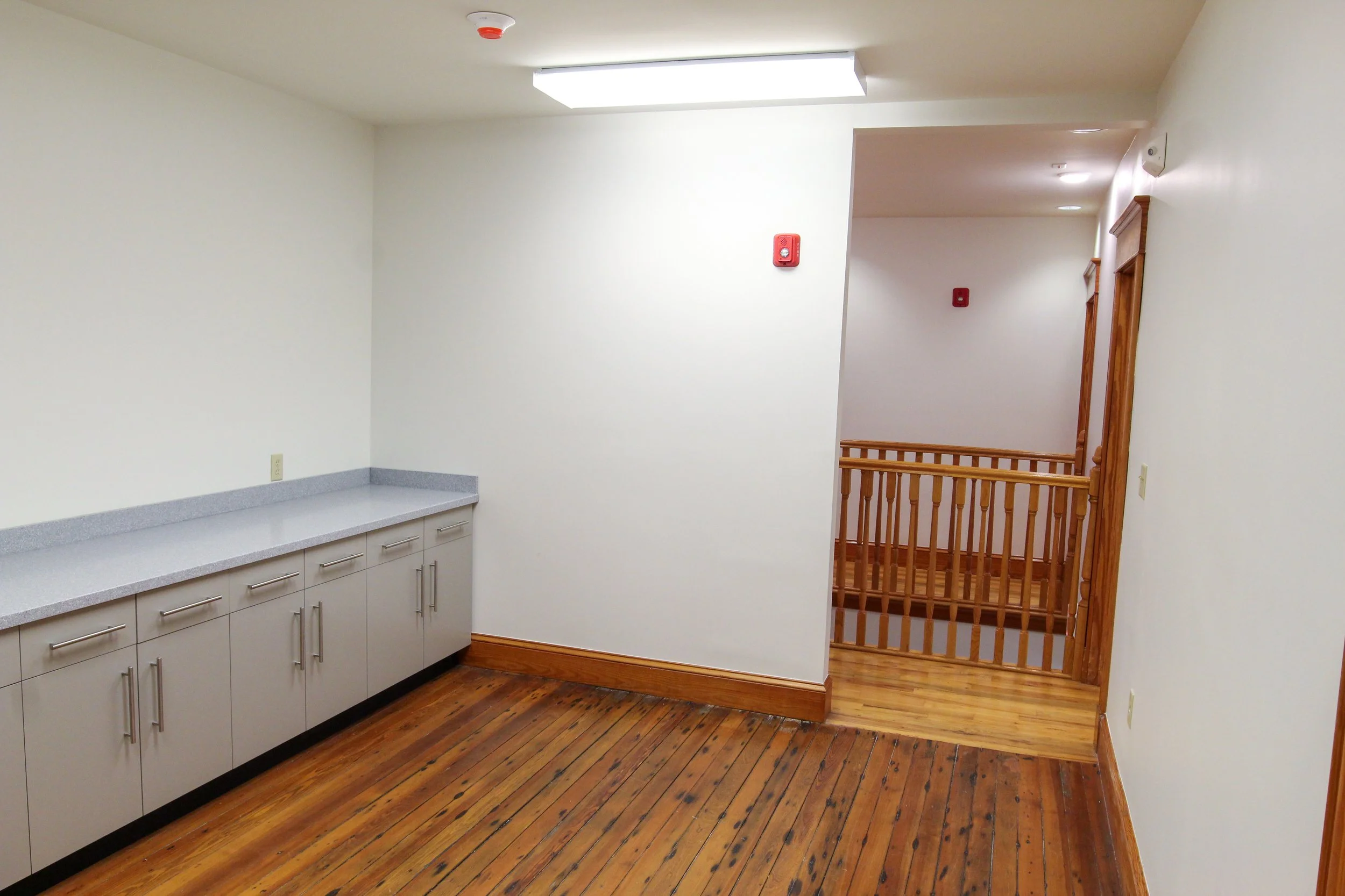 Empty room with a counter on the left, wooden flooring, white walls, wooden railing in the background, ceiling light, smoke detector, and a red fire alarm on the wall.