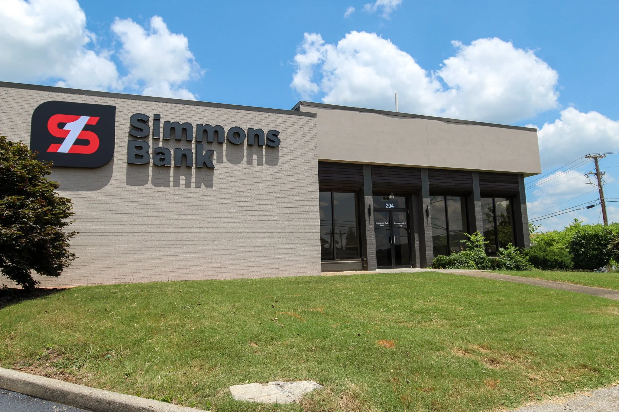 Exterior of a Simmons Bank branch with a logo on the left, a glass door labeled 204, and a green lawn in front, under a partly cloudy sky.