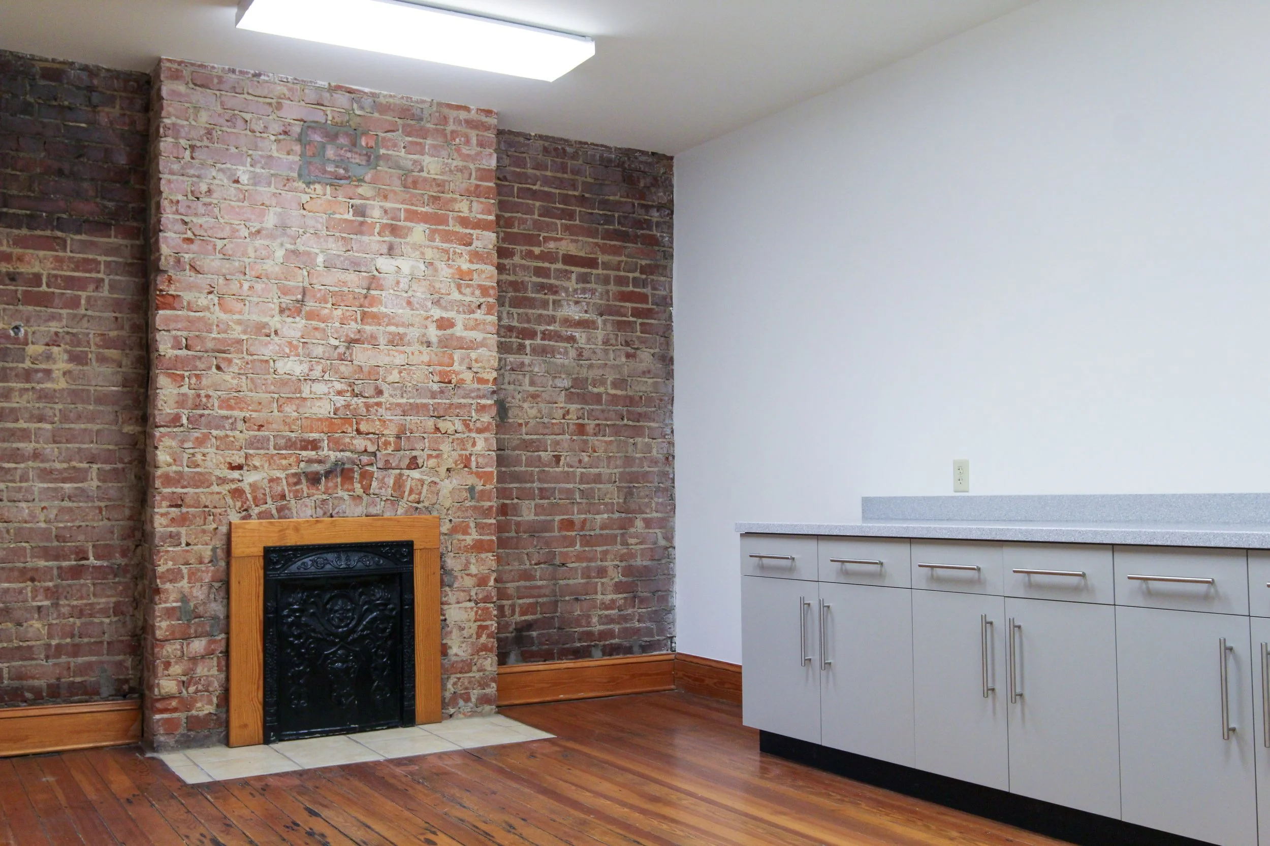Interior room with exposed brick wall, a small fireplace with a wooden mantel, and a white cabinet with a gray countertop against the right wall.