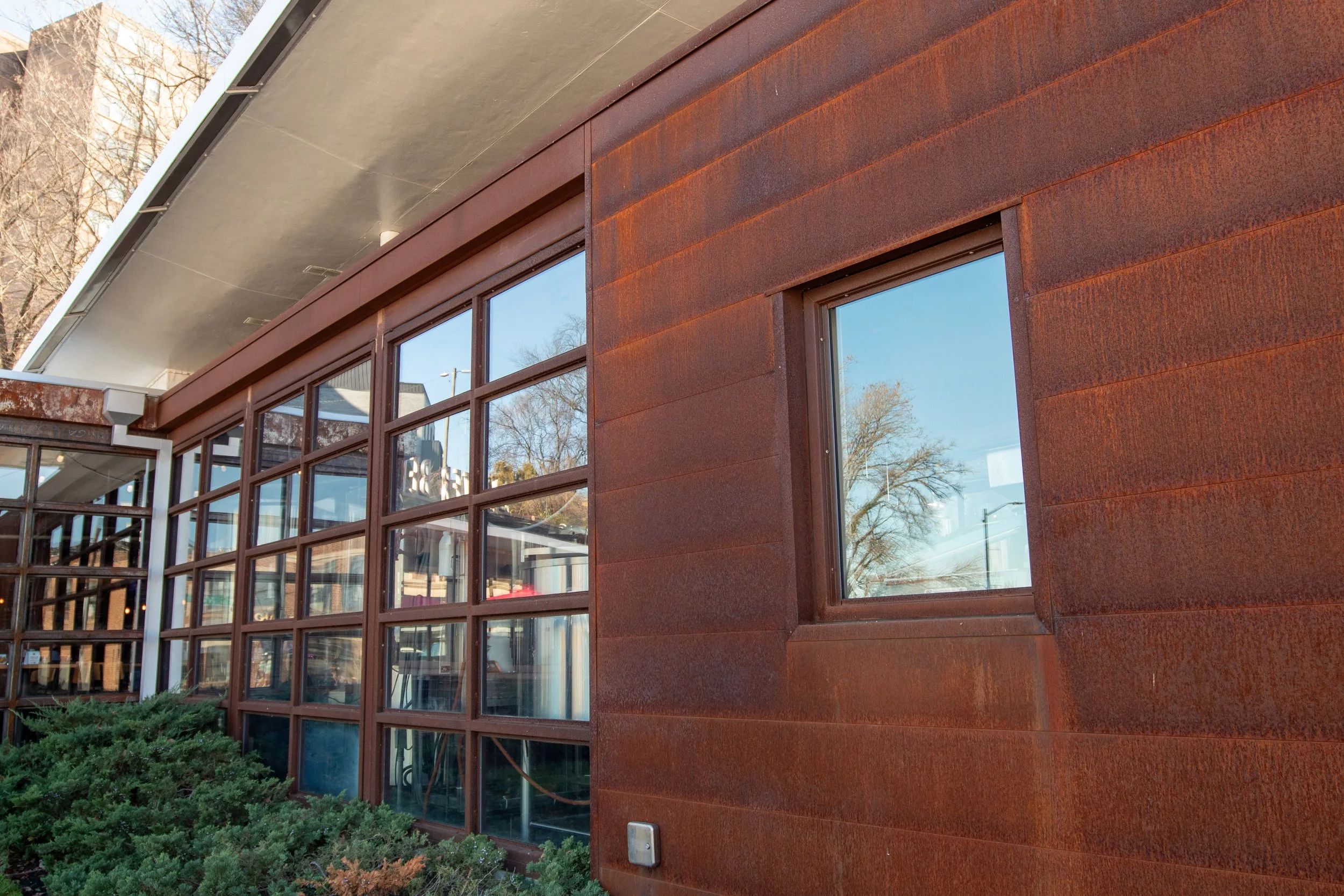 Close-up of a modern building with rust-colored metal siding and large windows, reflecting trees and the sky.