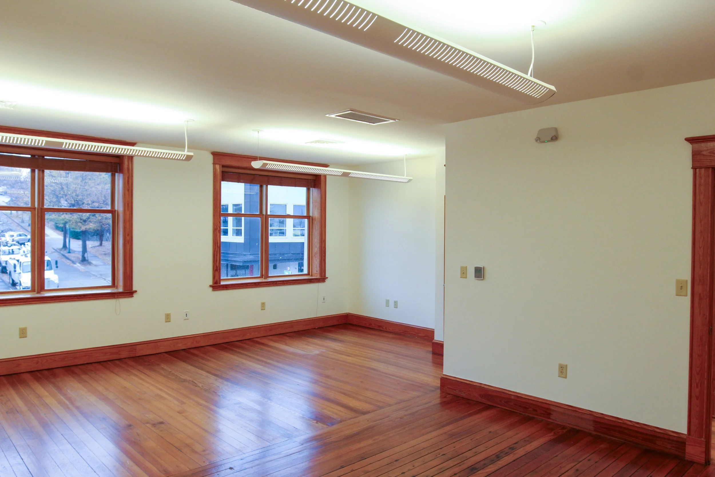 Empty room with hardwood floors, large windows, and white walls, with wooden trim around the windows and baseboards.