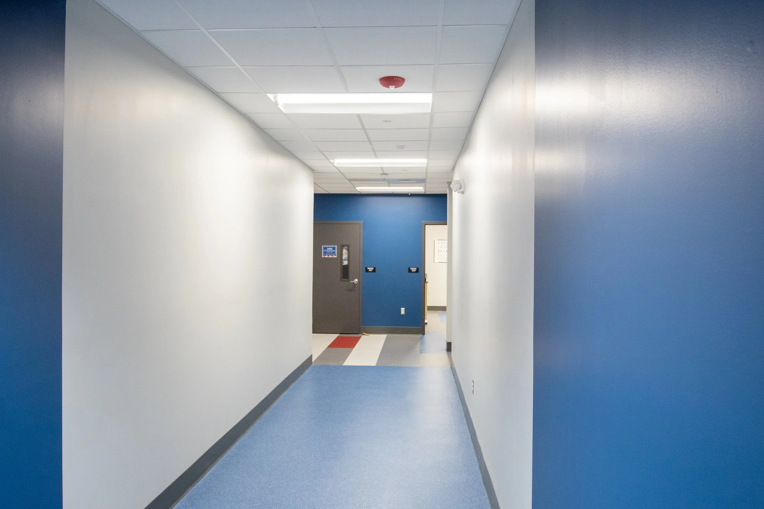 Empty hospital hallway with blue and white walls, blue flooring, and a gray door at the end.