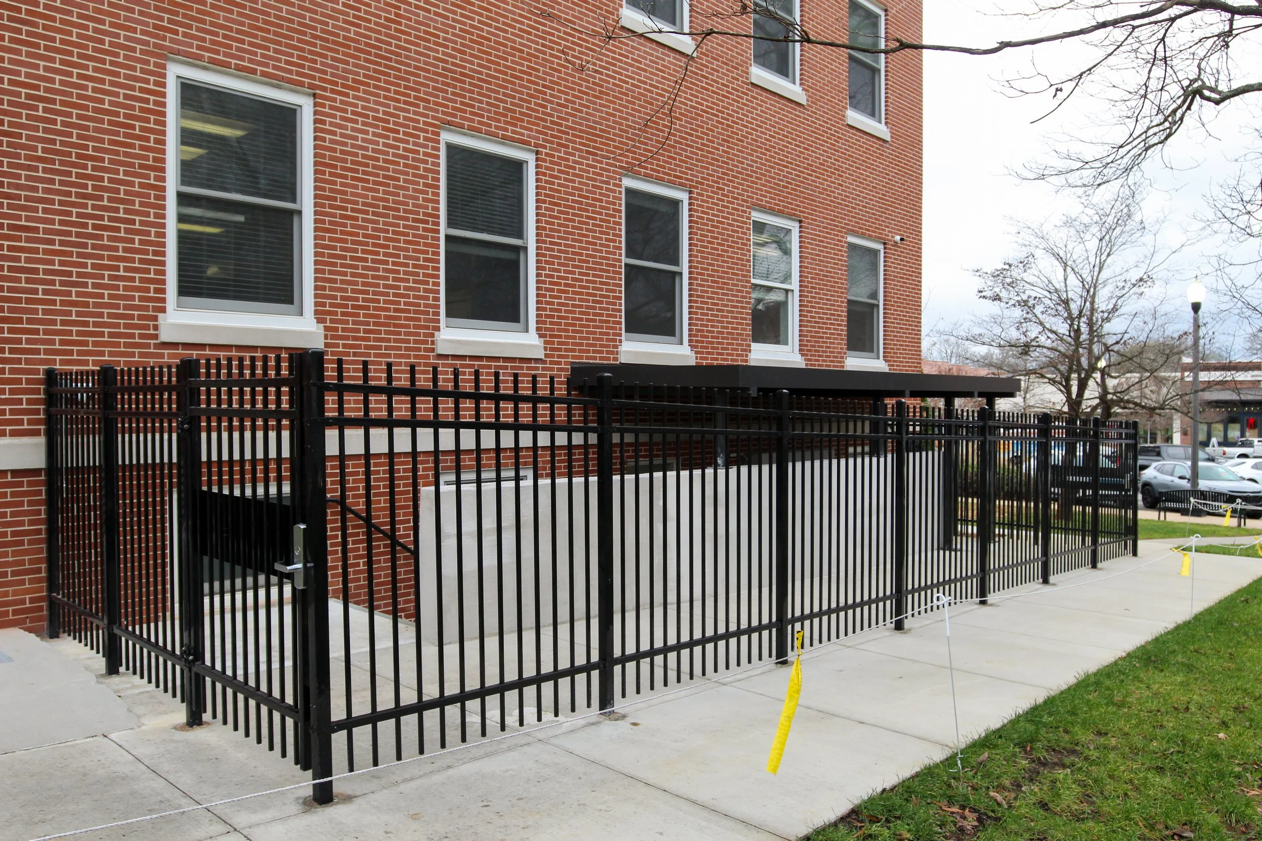 Black metal fence enclosing a concrete ramp and stairs leading to a multi-story brick building with several windows and a small overhang. There are trees and parked cars in the background.