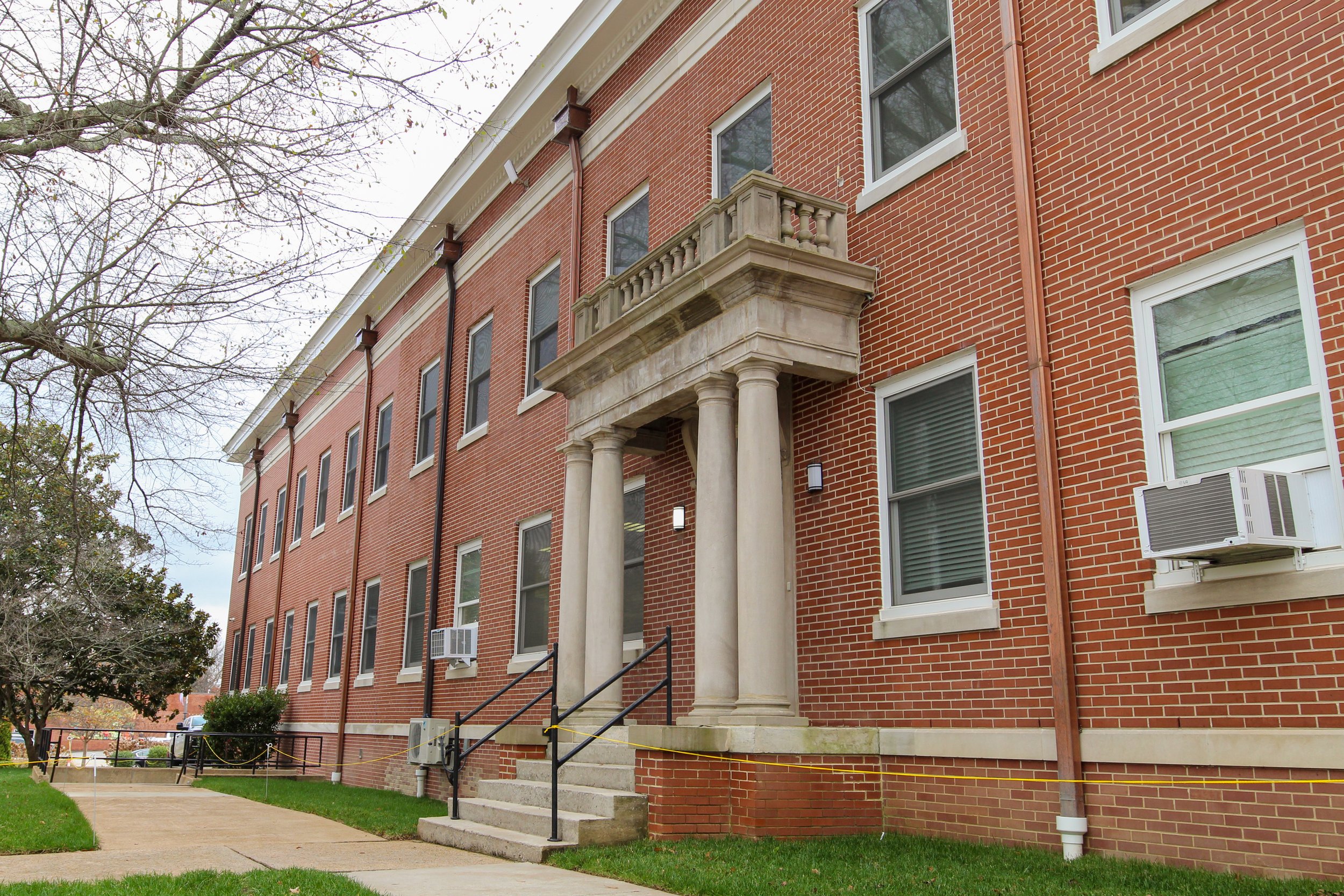 A red brick apartment building with a classical portico supported by two columns, window air conditioning units, stairs leading up to the entrance, and a lawn with a sidewalk.