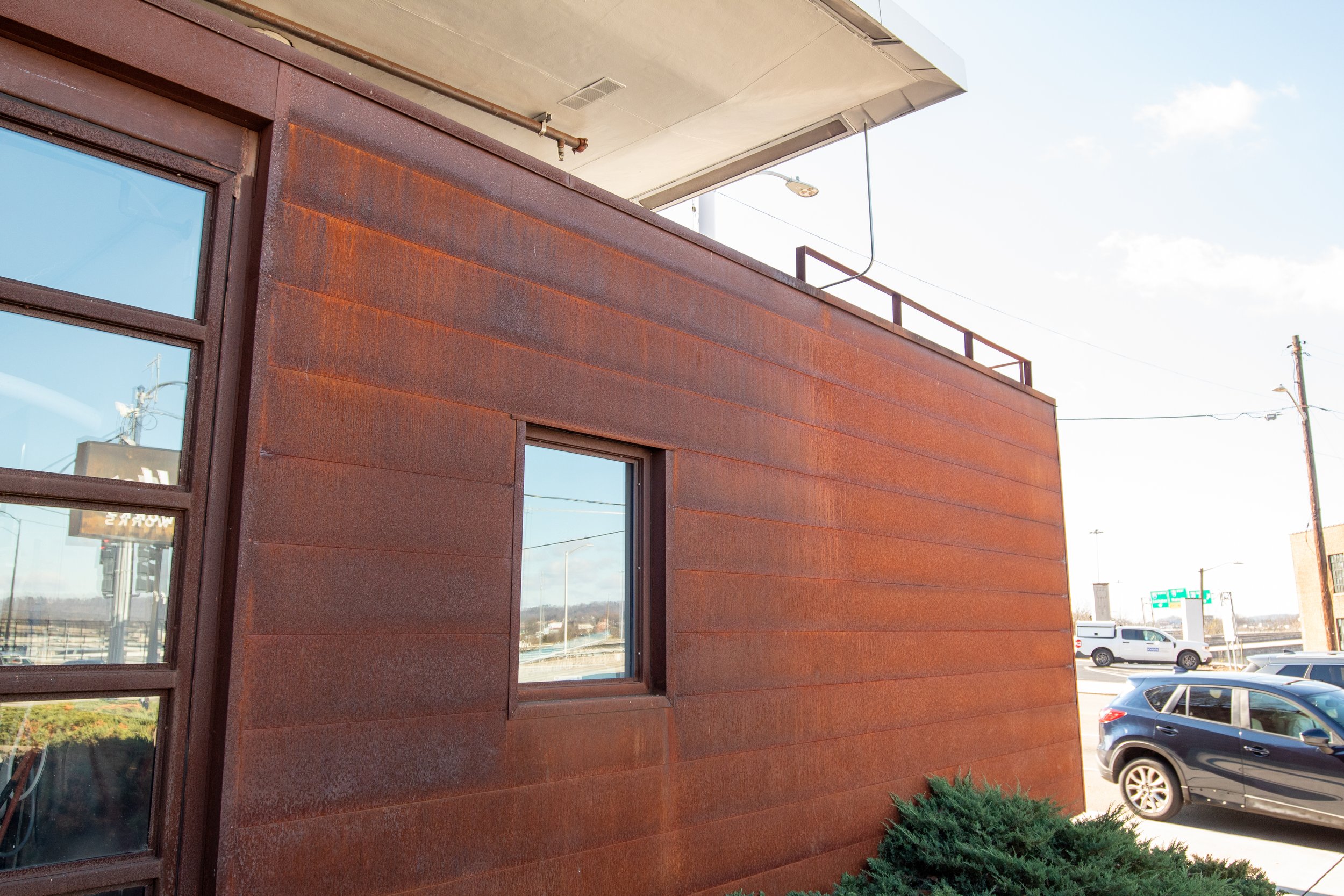 Close-up of a modern building with a rusted metal exterior, a small window, and a corner of a glass door visible on the left. There are cars and utility poles in the background under a clear sky.