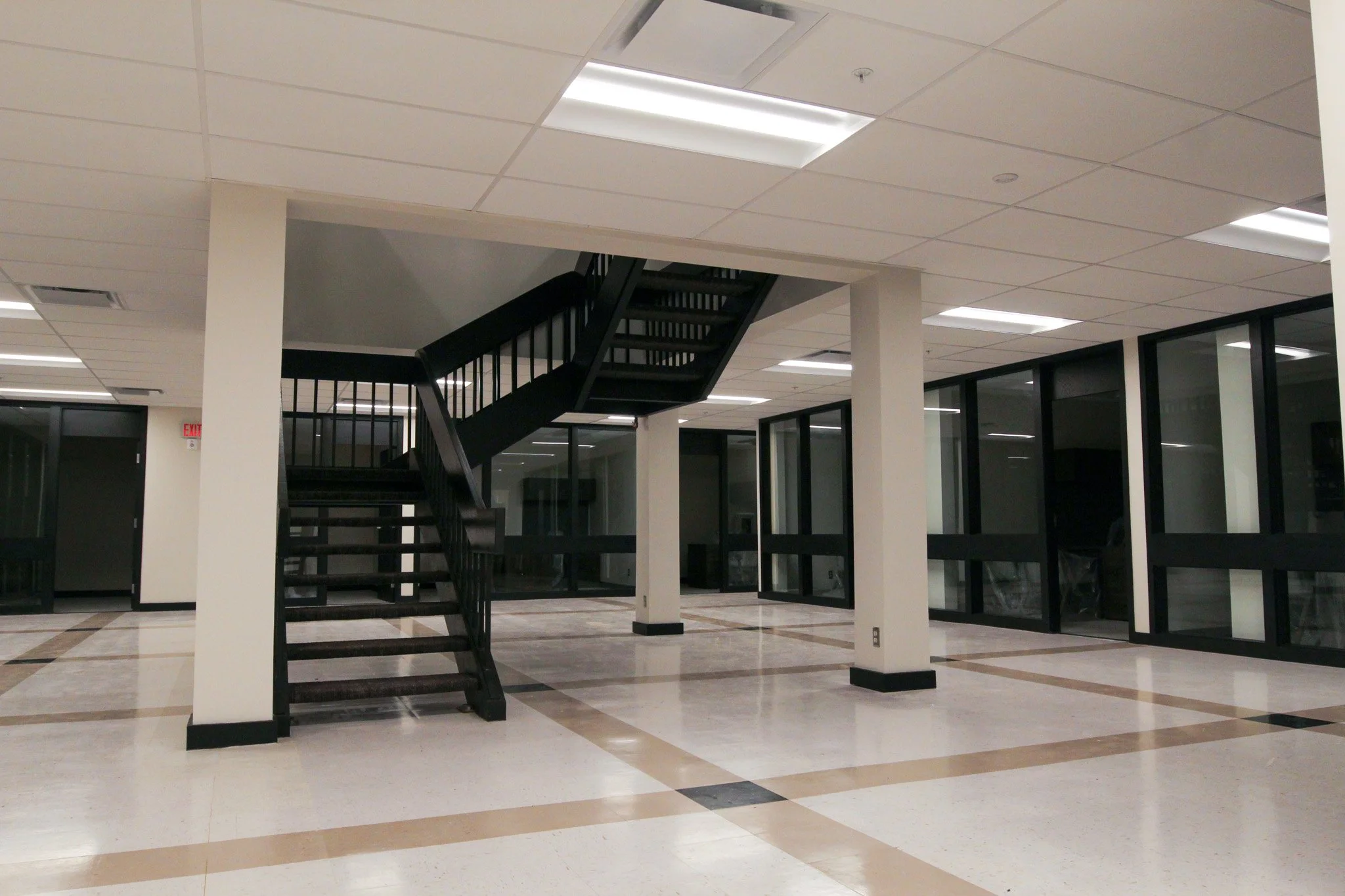 Interior of a building with an open area featuring a black staircase and glass doors, with white ceiling tiles and fluorescent lighting.