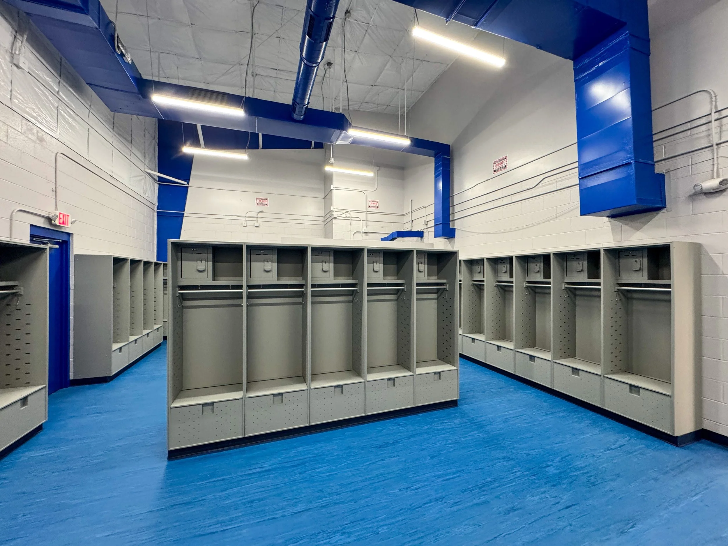 Empty gym storage lockers with gray lockers, blue floors, and white walls. Blue industrial ventilation ducts run along the ceiling, and there are exit signs on the walls.