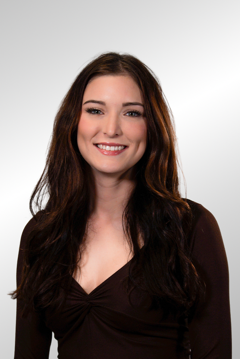 A young woman with long, wavy brown hair smiling at the camera. She is wearing a black top with a tan, pinstriped vest and has her arms crossed.