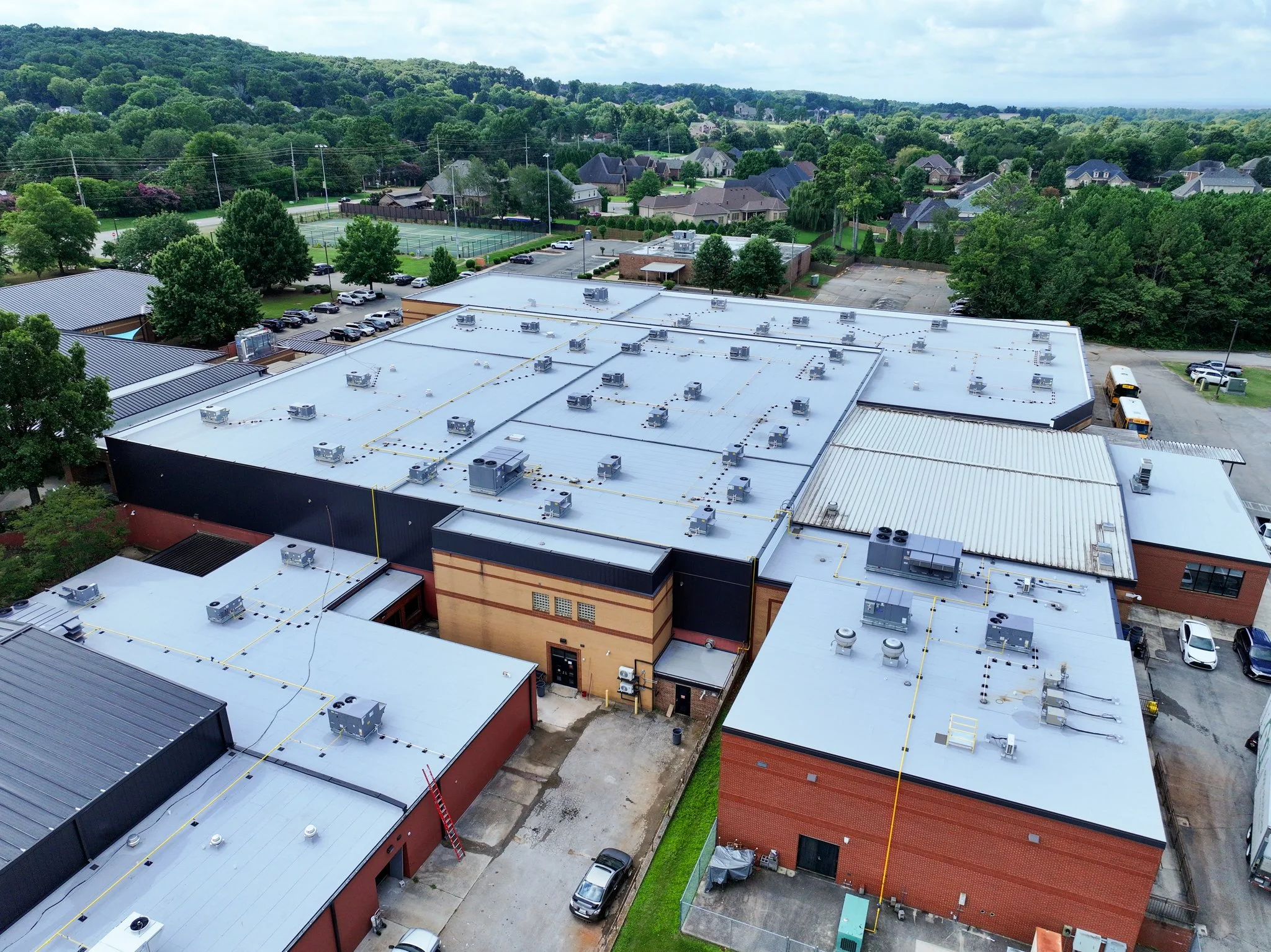 An aerial view of Discovery Middle School with a white flat roof equipped with numerous air conditioning units. Surrounding the building are parking lots, green trees, and residential houses in the background, with a tennis court nearby.