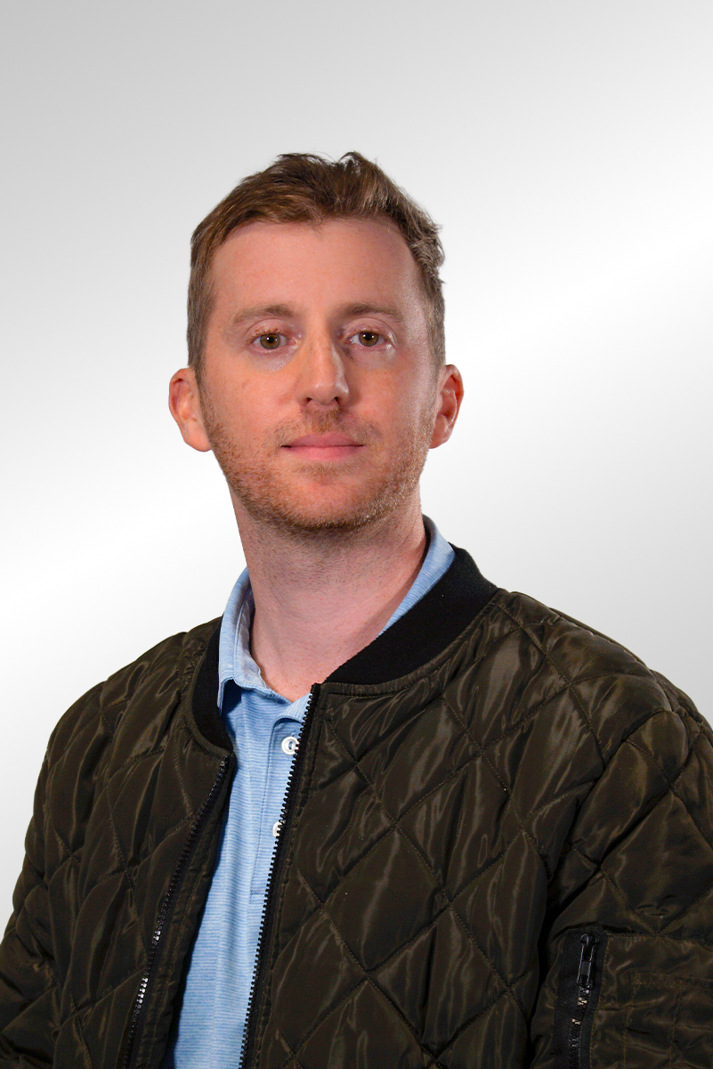 A smiling man with short light brown hair and a beard wearing a light striped polo shirt against a dark background.