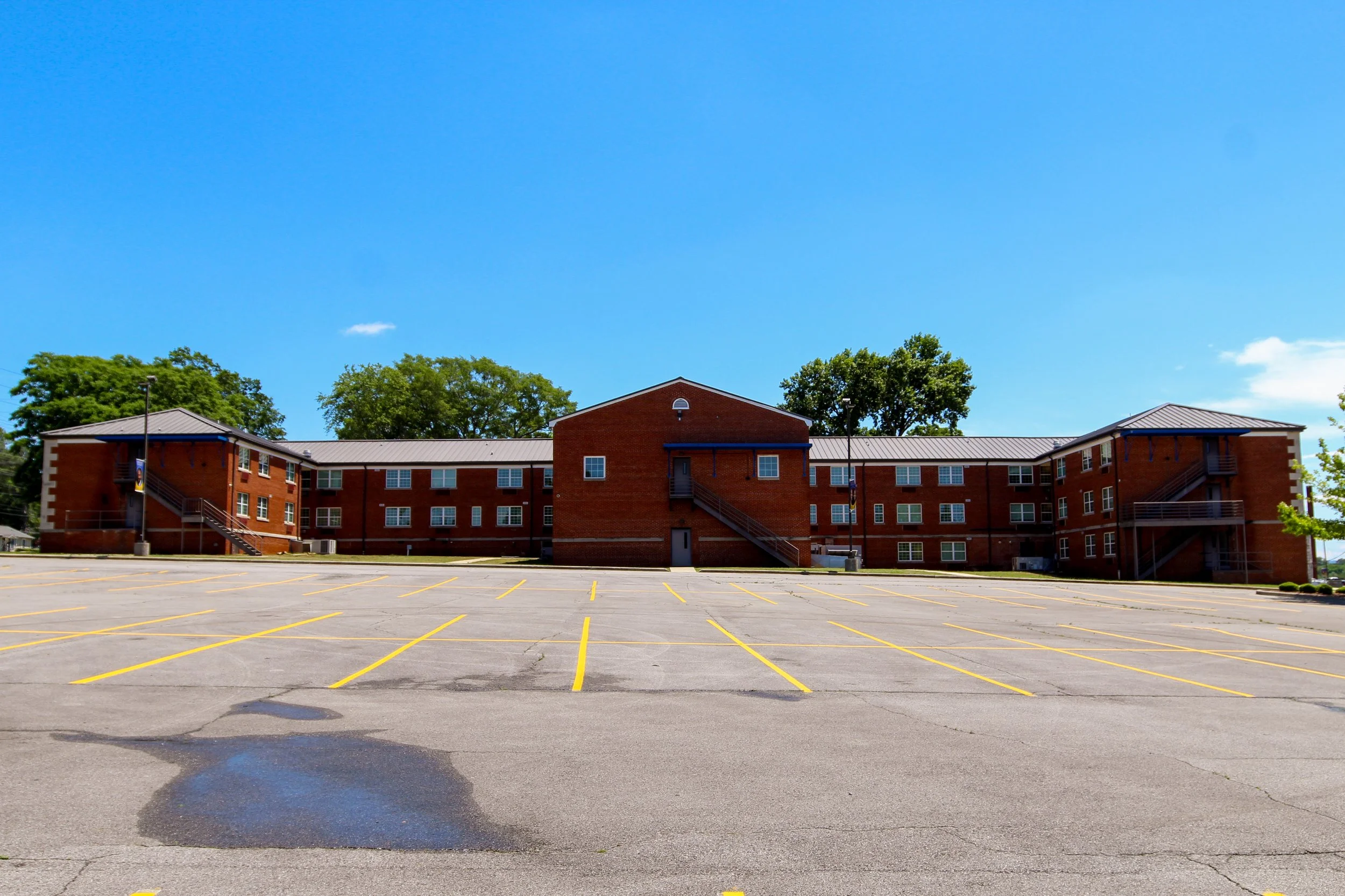 A large brick apartment building under a bright blue sky with a few clouds. The foreground shows an empty parking lot with yellow parking lines and some cracks in the pavement. Historic Remodeled by P&C Construction, Inc. - Chattanooga, Tn.