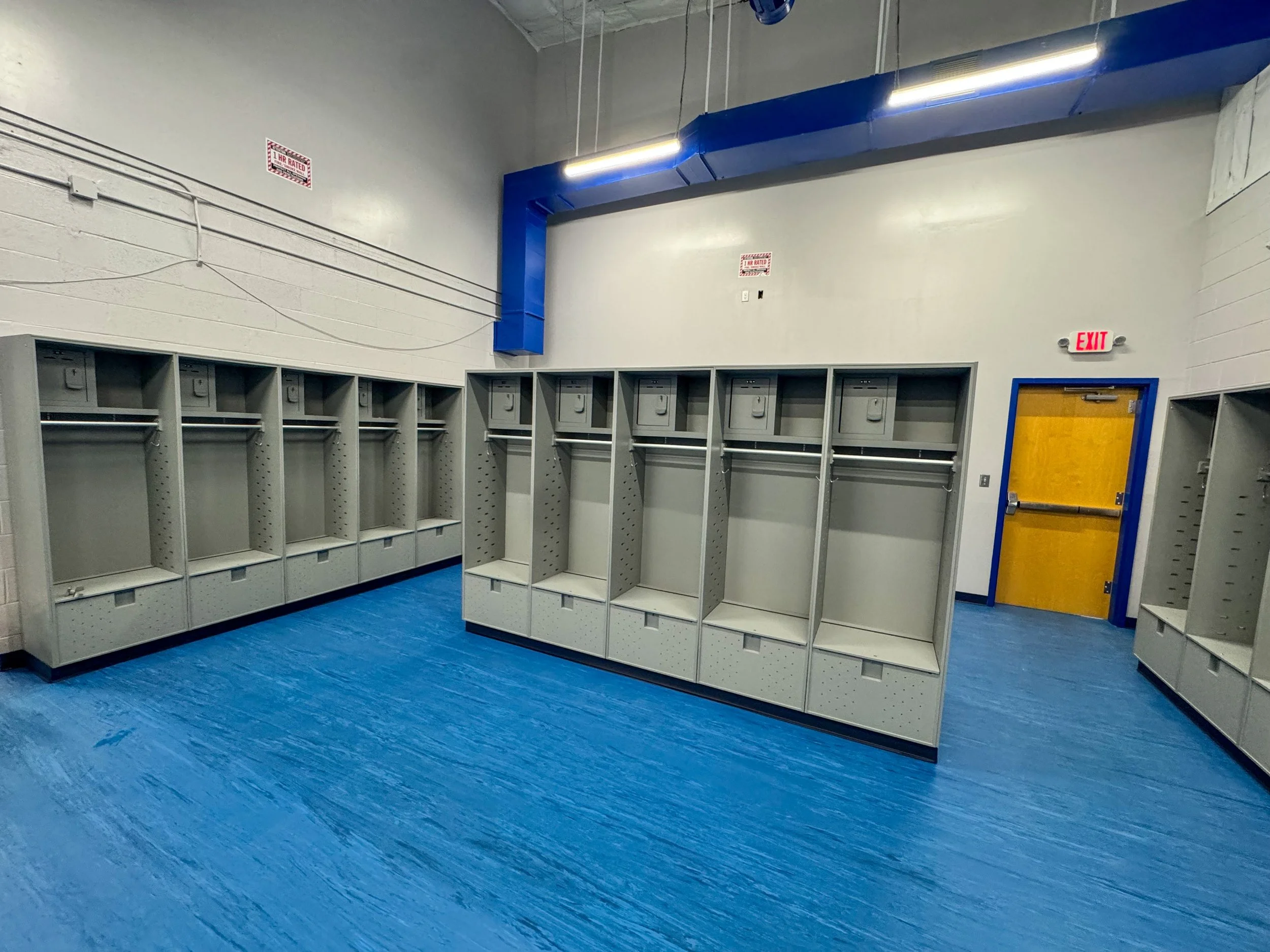 Empty locker room with rows of gray lockers, blue flooring, a yellow door, and an exit sign.