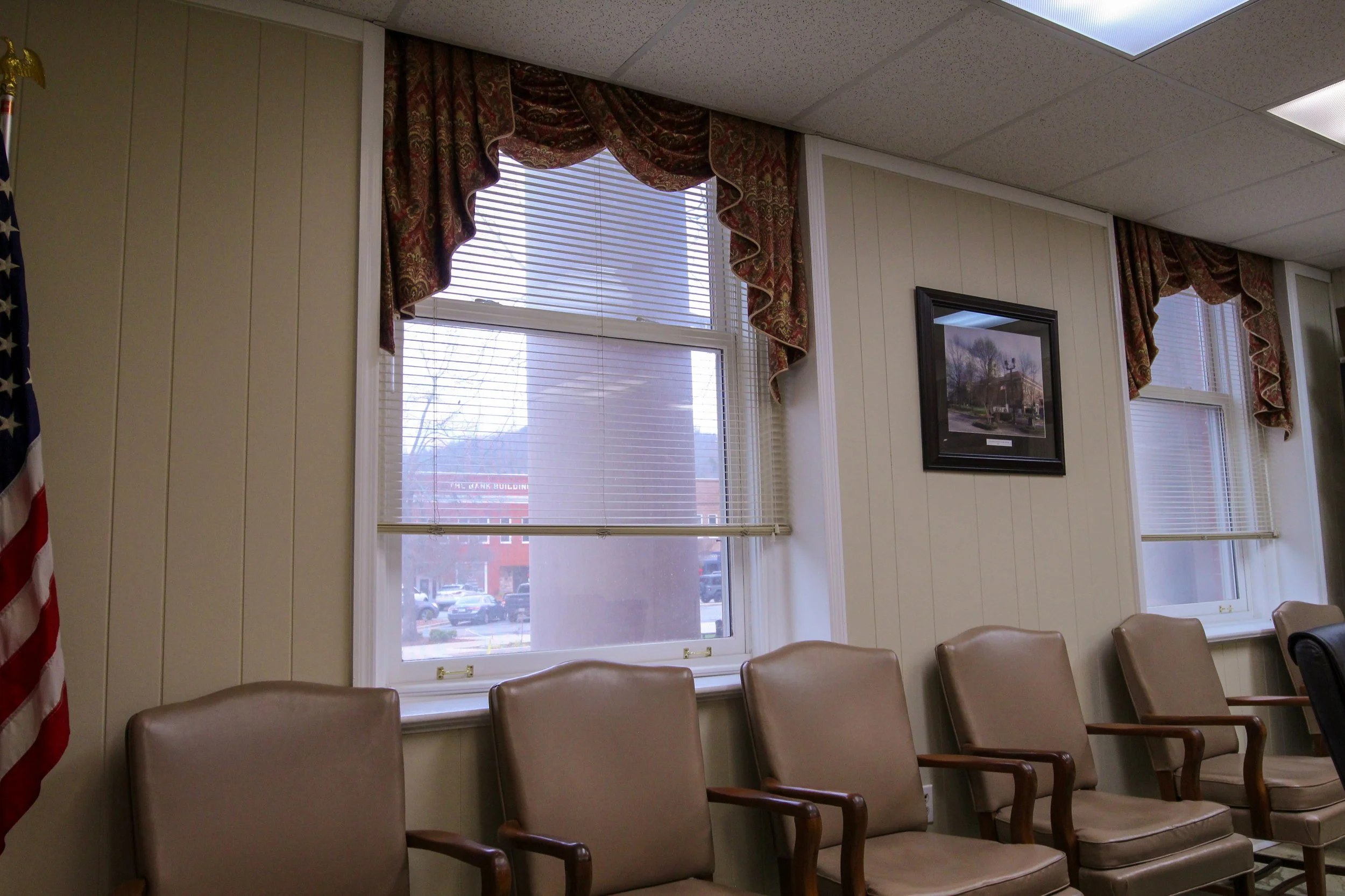 A room with beige chairs arranged in a row, white walls with vertical paneling, three windows with blinds and patterned drapes, a framed picture on the wall, and part of an American flag visible on the left.