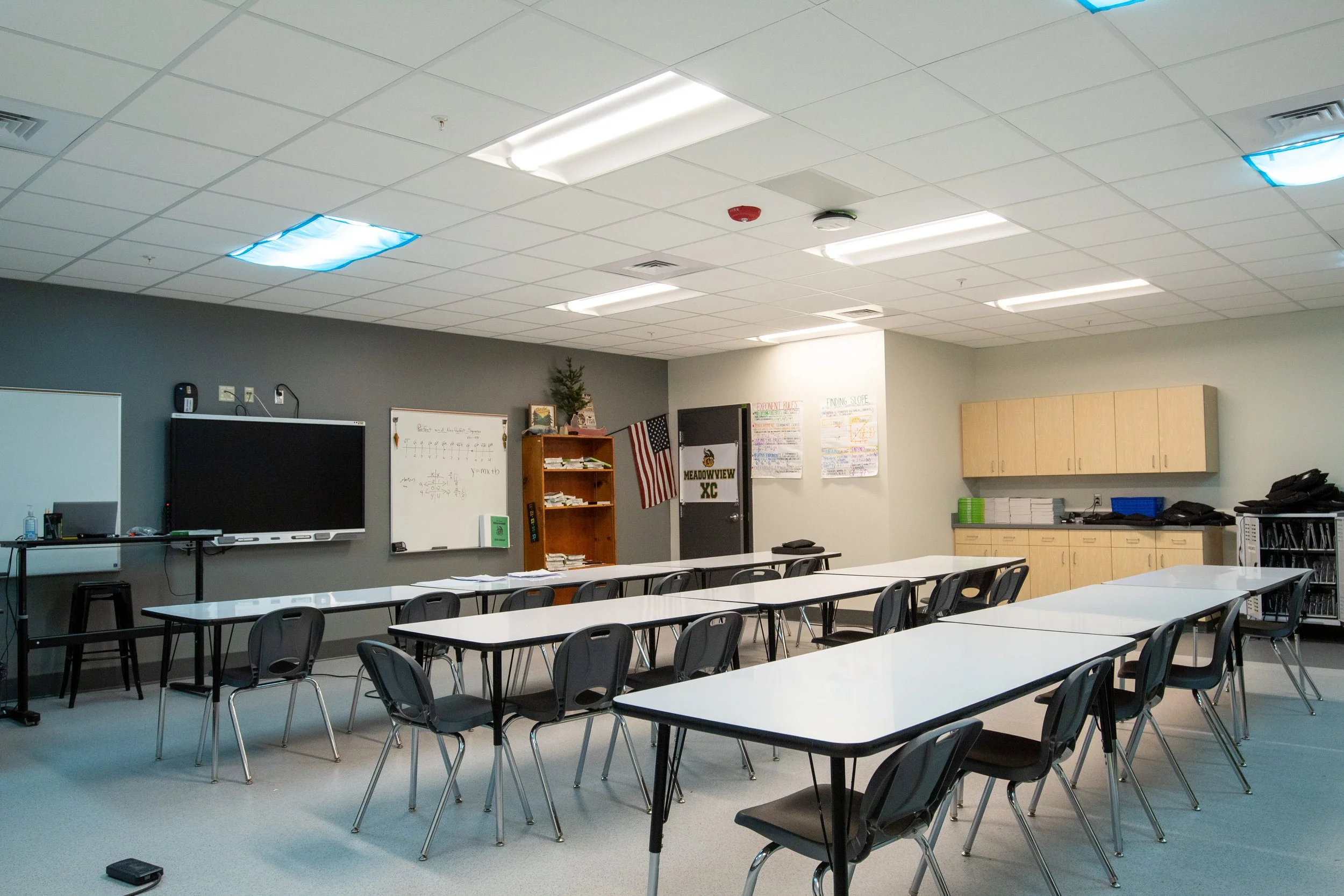 Empty classroom with tables and chairs, classroom supplies, a whiteboard, an interactive display, and posters on the wall.