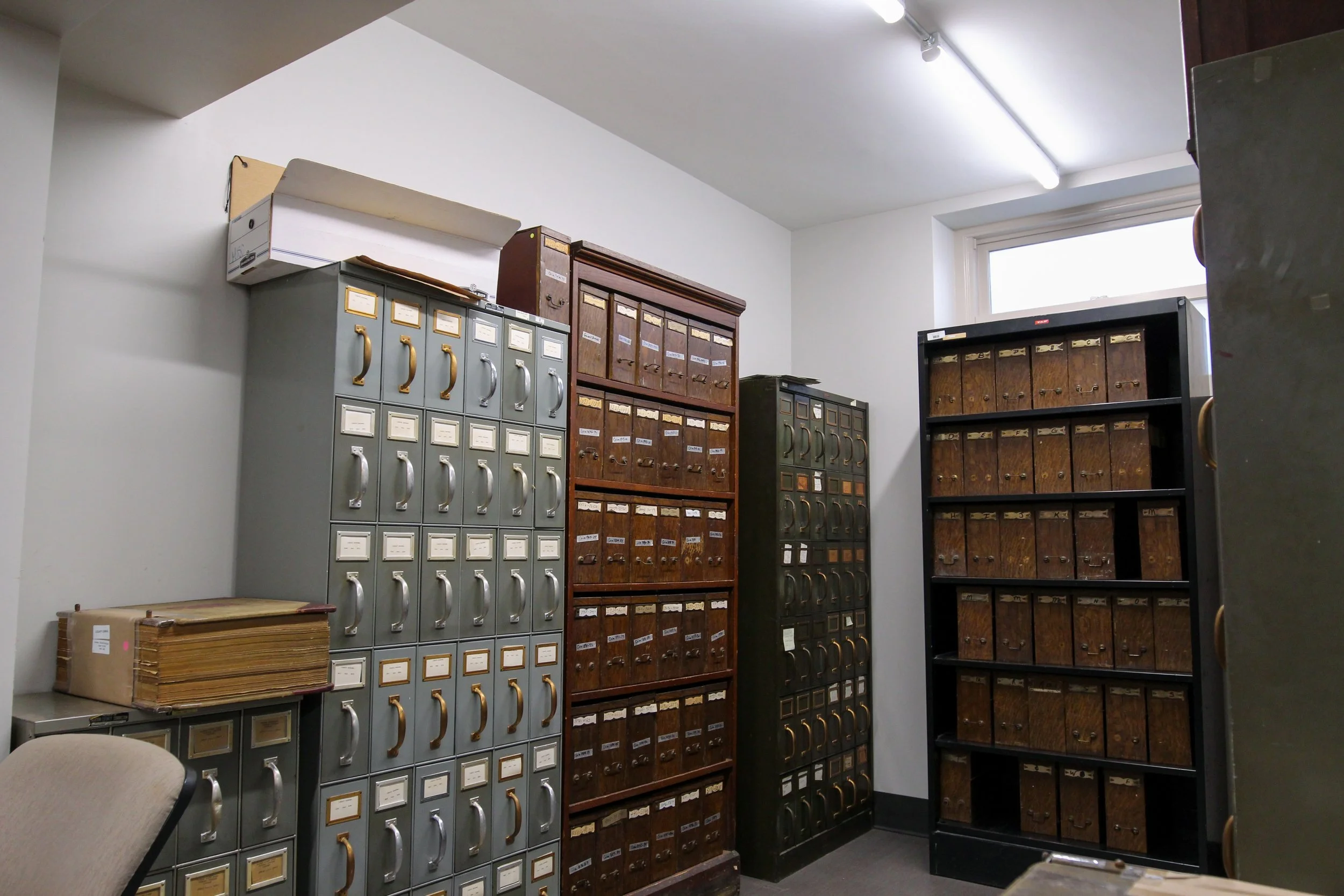 A room with metal and wooden filing cabinets filled with files and labels, a stack of folders on a small table, a beige office chair, and a window with a fluorescent light fixture overhead.