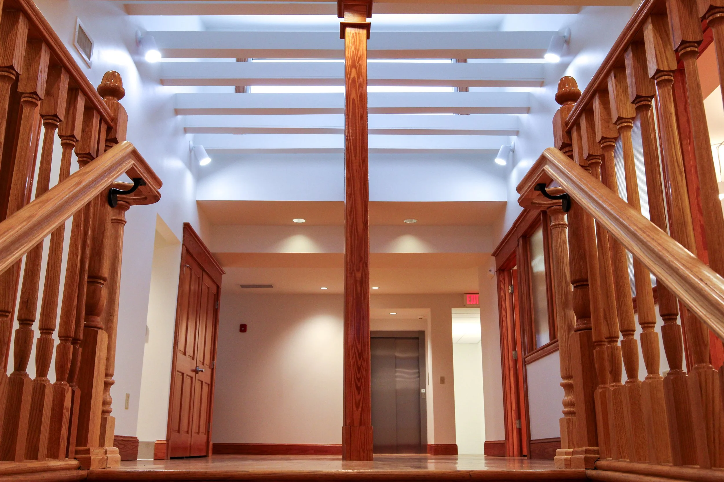 Interior view of a building with wooden stair railings, a central wooden column, and an elevator at the far end.