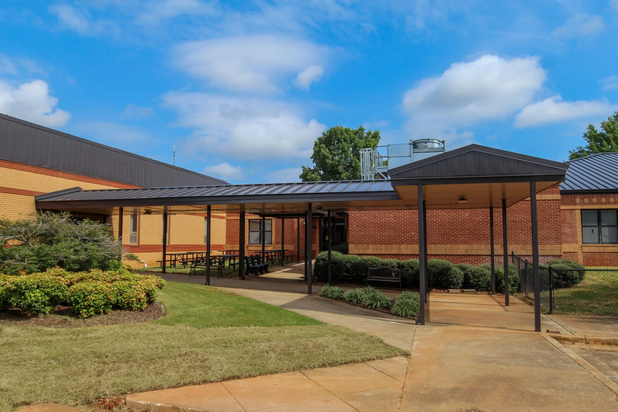 A school building with a covered outdoor seating area, landscaped yard, and a blue sky with clouds.