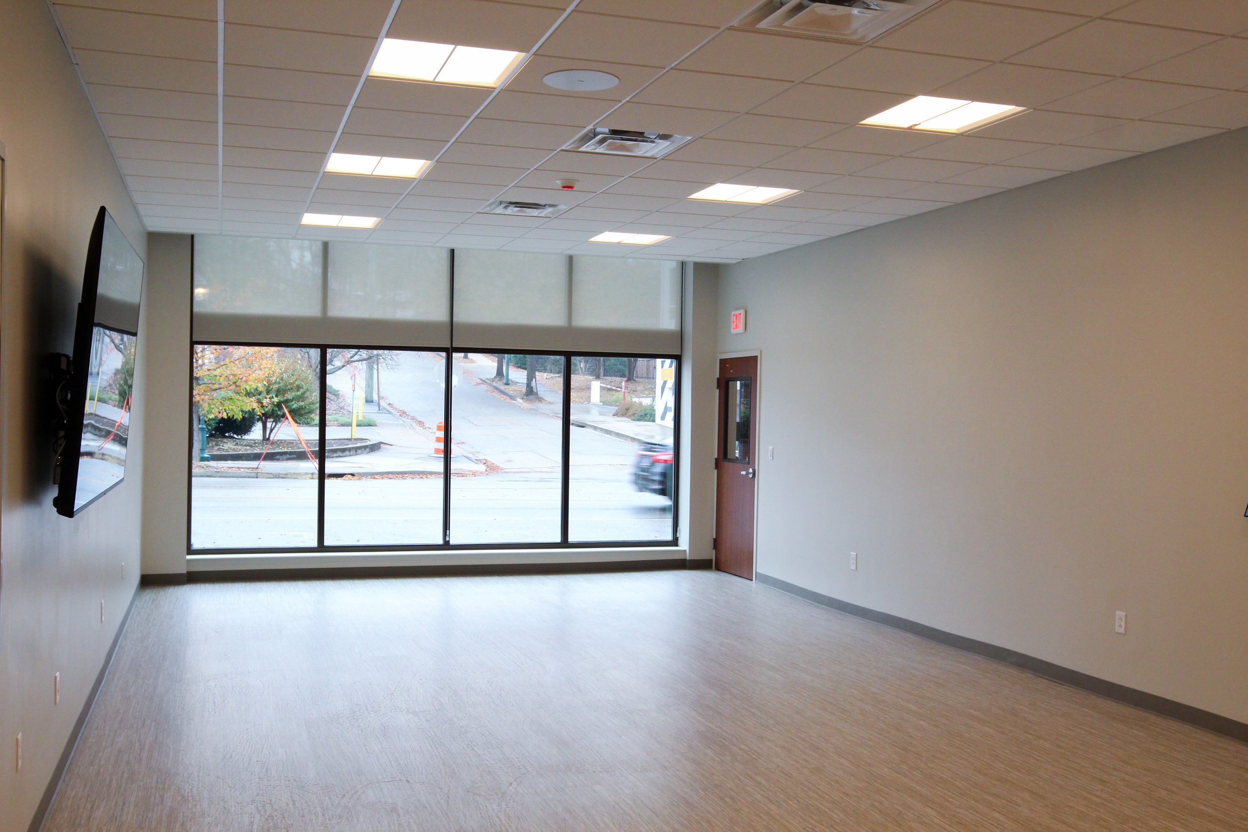 Empty office or conference room with large windows, a wall-mounted TV, light-colored carpet, and a ceiling with fluorescent lights. Outside view includes a street with a car passing by, trees, and construction cones.