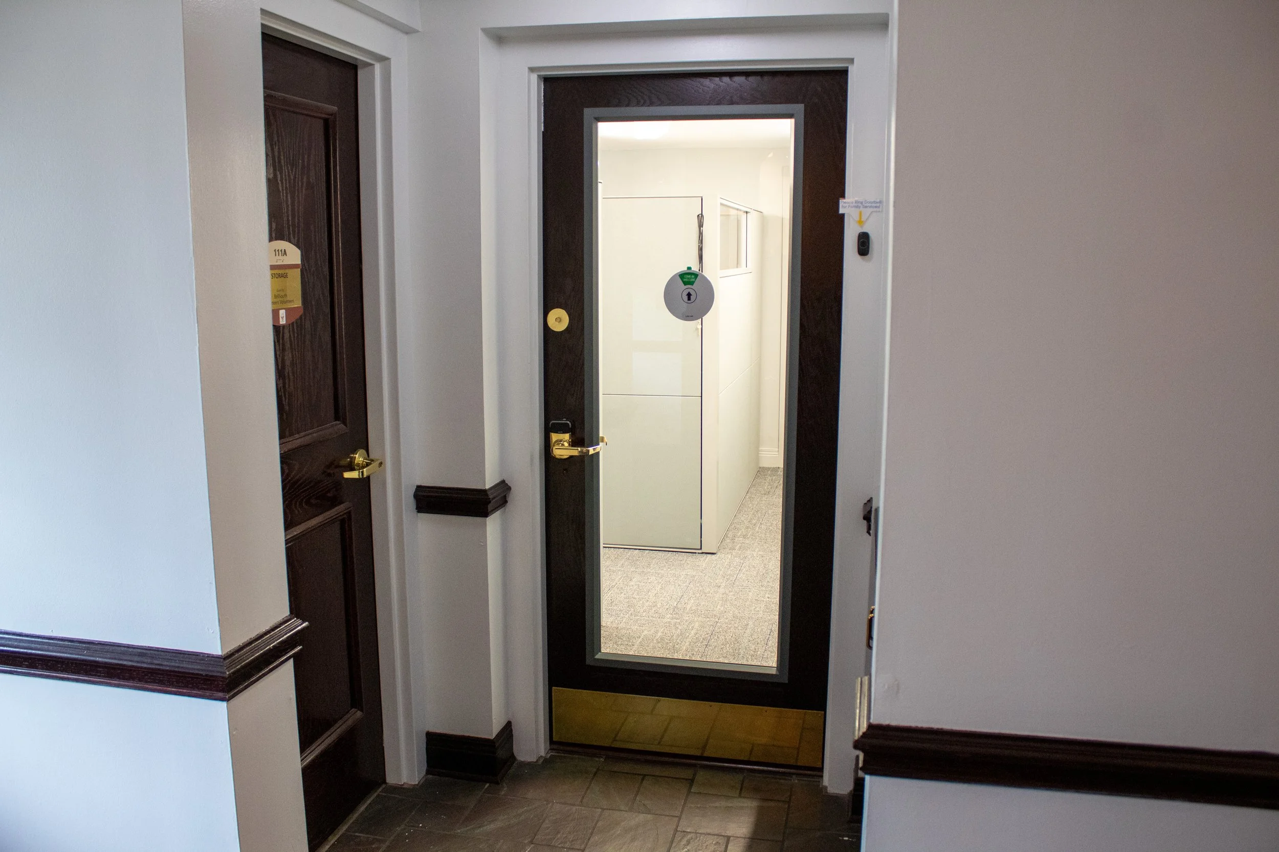 An interior hallway with two closed wood doors, one with a gold handle, and a black-framed glass door with a lock and a security barcode scanner. The hallway has white walls with dark wood trim and a tiled floor.