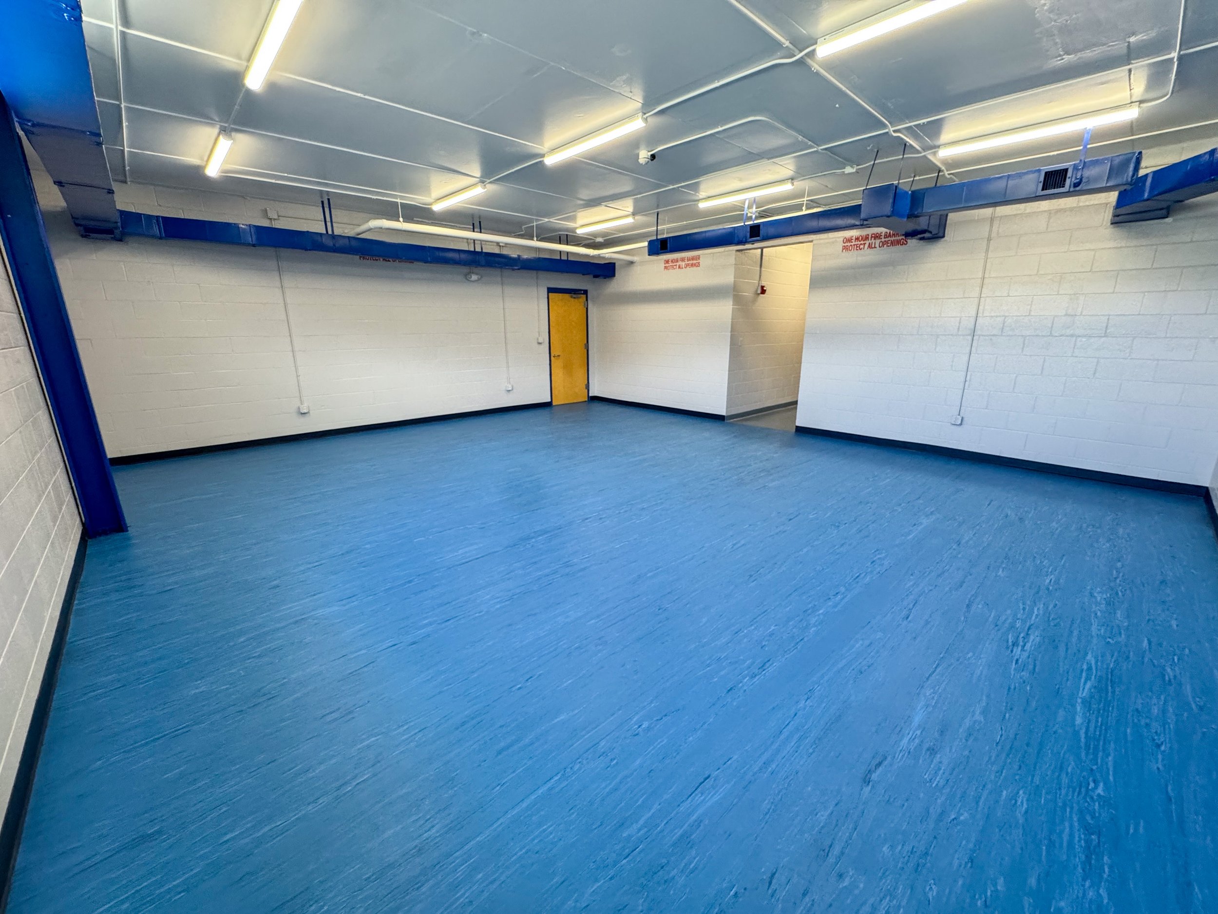 Empty room with white brick walls, blue floor, exposed pipes and ductwork, fluorescent lighting, and a yellow door.
