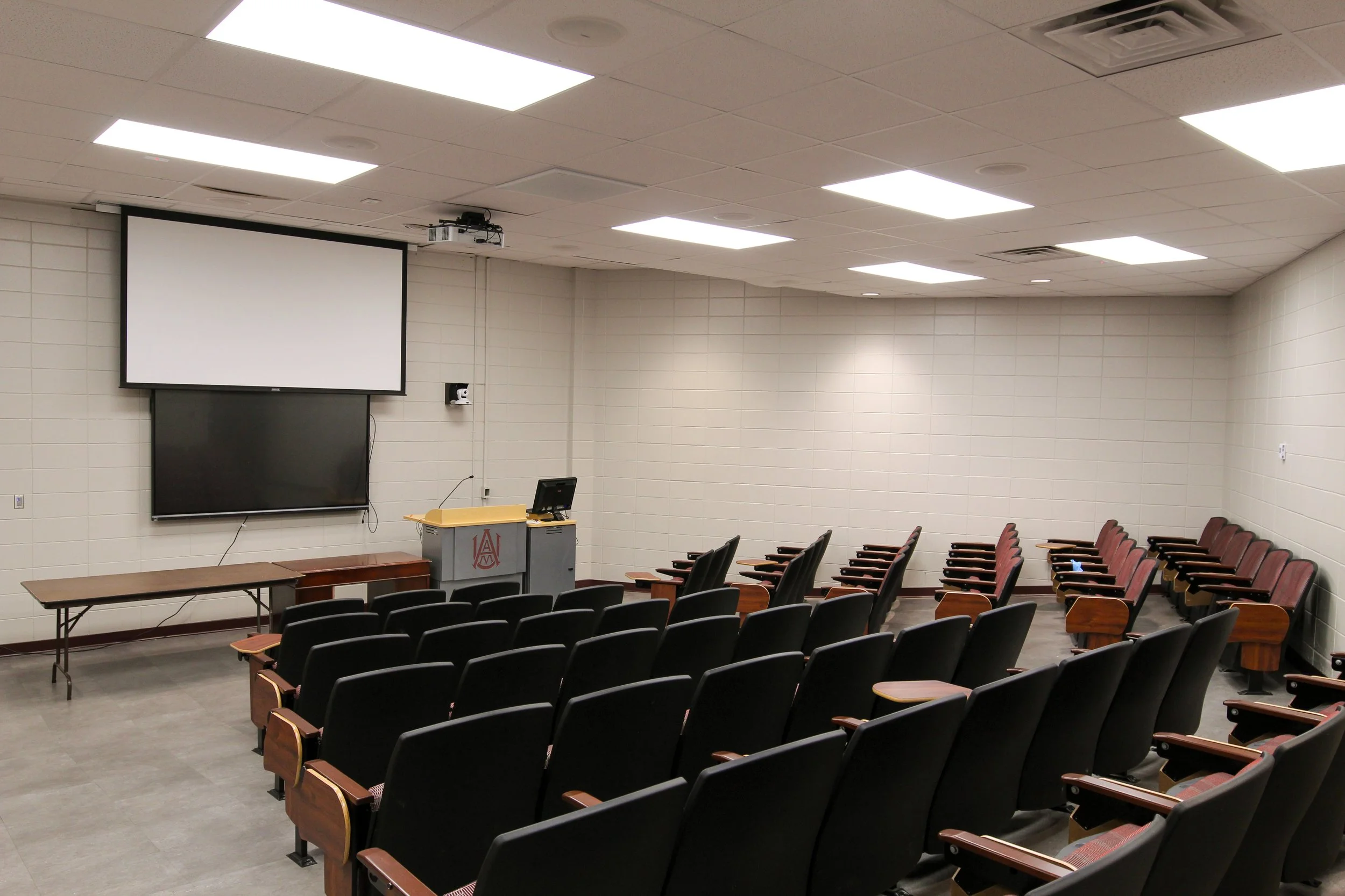 Empty classroom or lecture hall with rows of black and maroon cushioned chairs facing a projection screen and a blackboard, with a wooden podium and a television, all on a tiled floor.