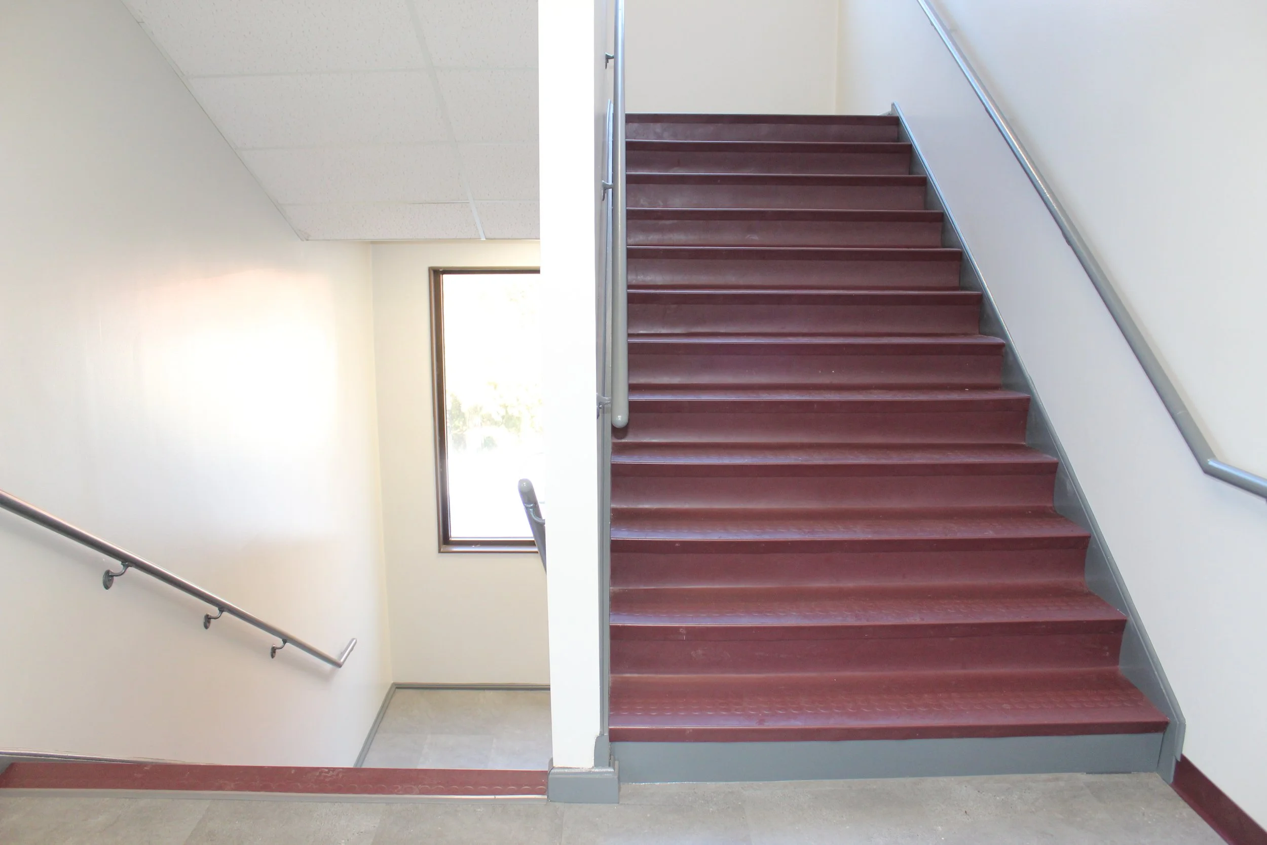 Interior of a staircase with red steps and metal handrails, with a window illuminating the area.