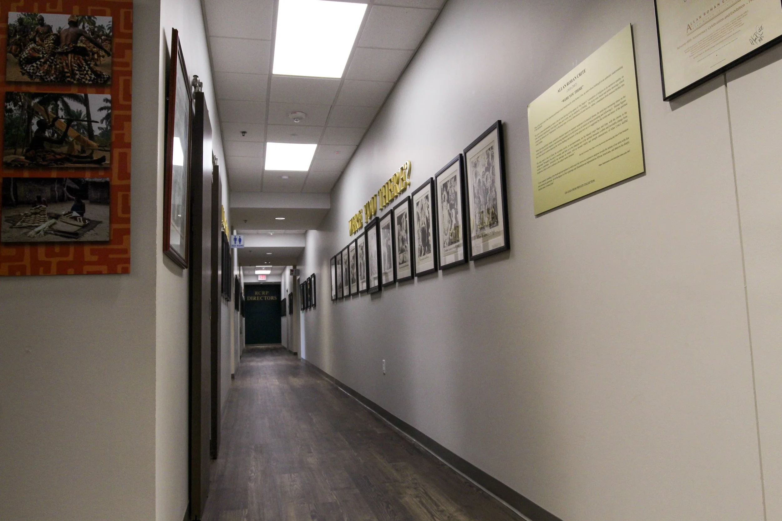 A hallway in a building with framed artwork and yellow signs on the wall, and a sign indicating the restrooms at the end. Design build by P&C Construction, Inc. - Chattanooga, Tn.