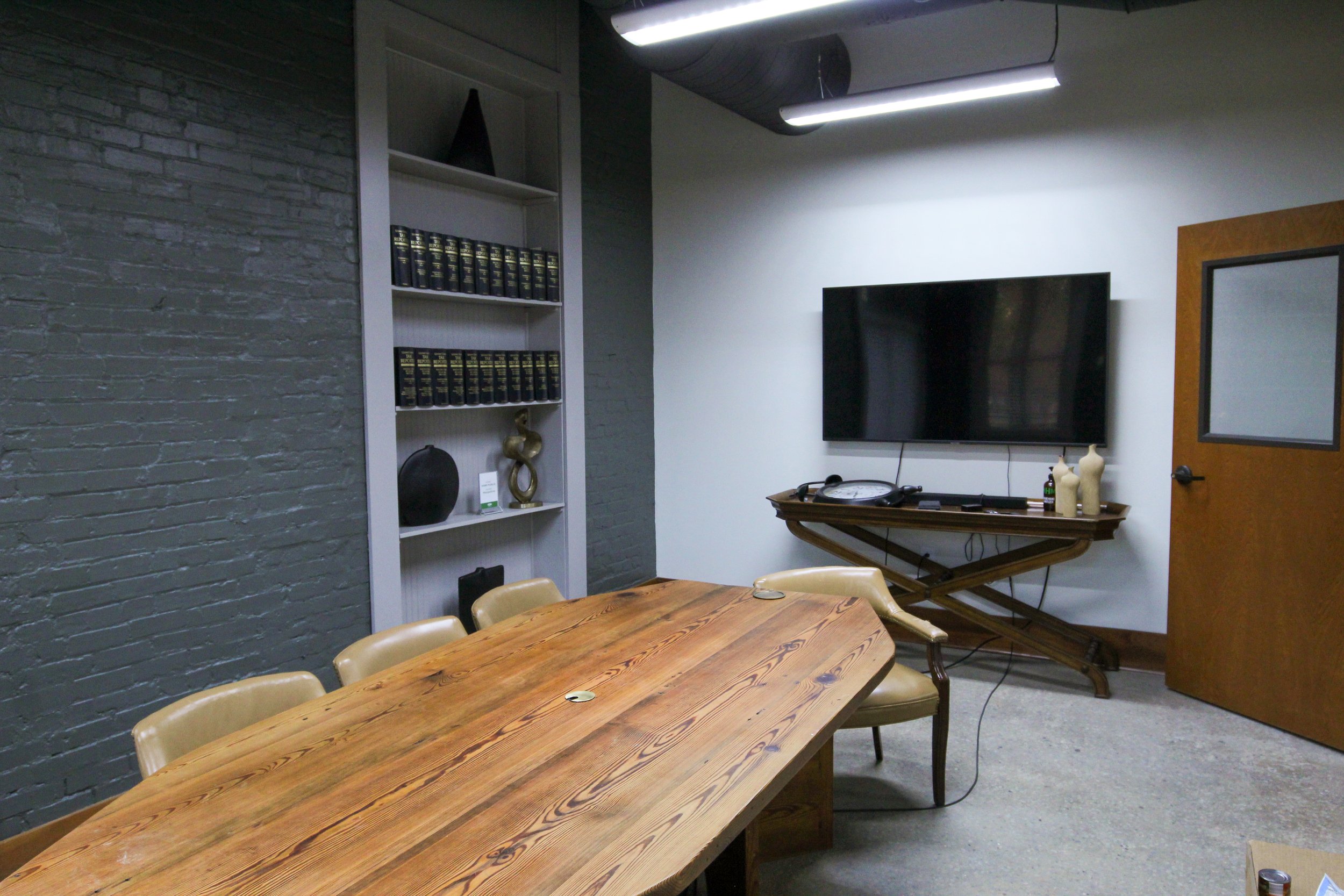 An empty conference room with a wooden table and beige chairs, a large flat-screen TV on the wall, a door, a white bookshelf with decor items and books, and a table with bottles, featuring a gray brick wall and a dark ceiling.