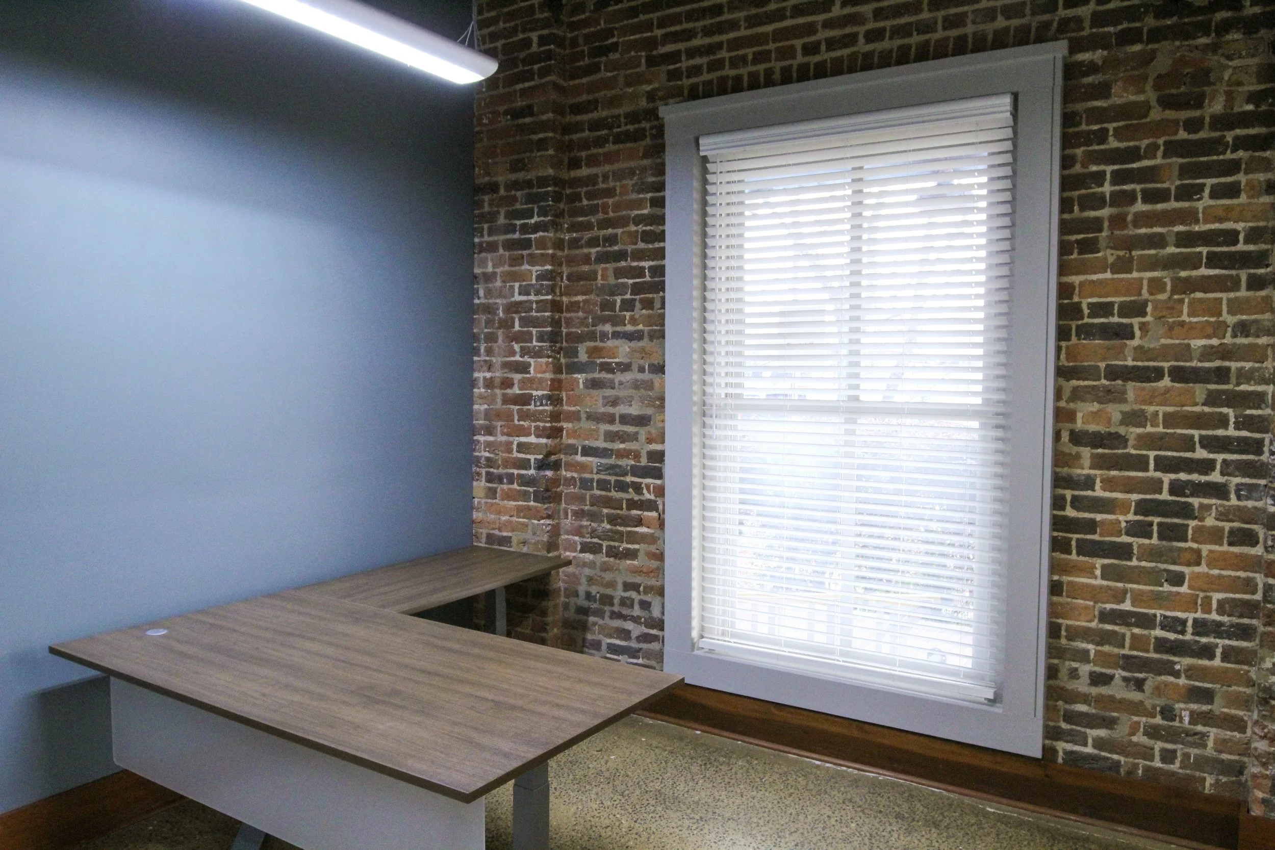 An empty office corner with a brick wall, a window with white blinds, a light blue wall, and a wooden L-shaped desk.
