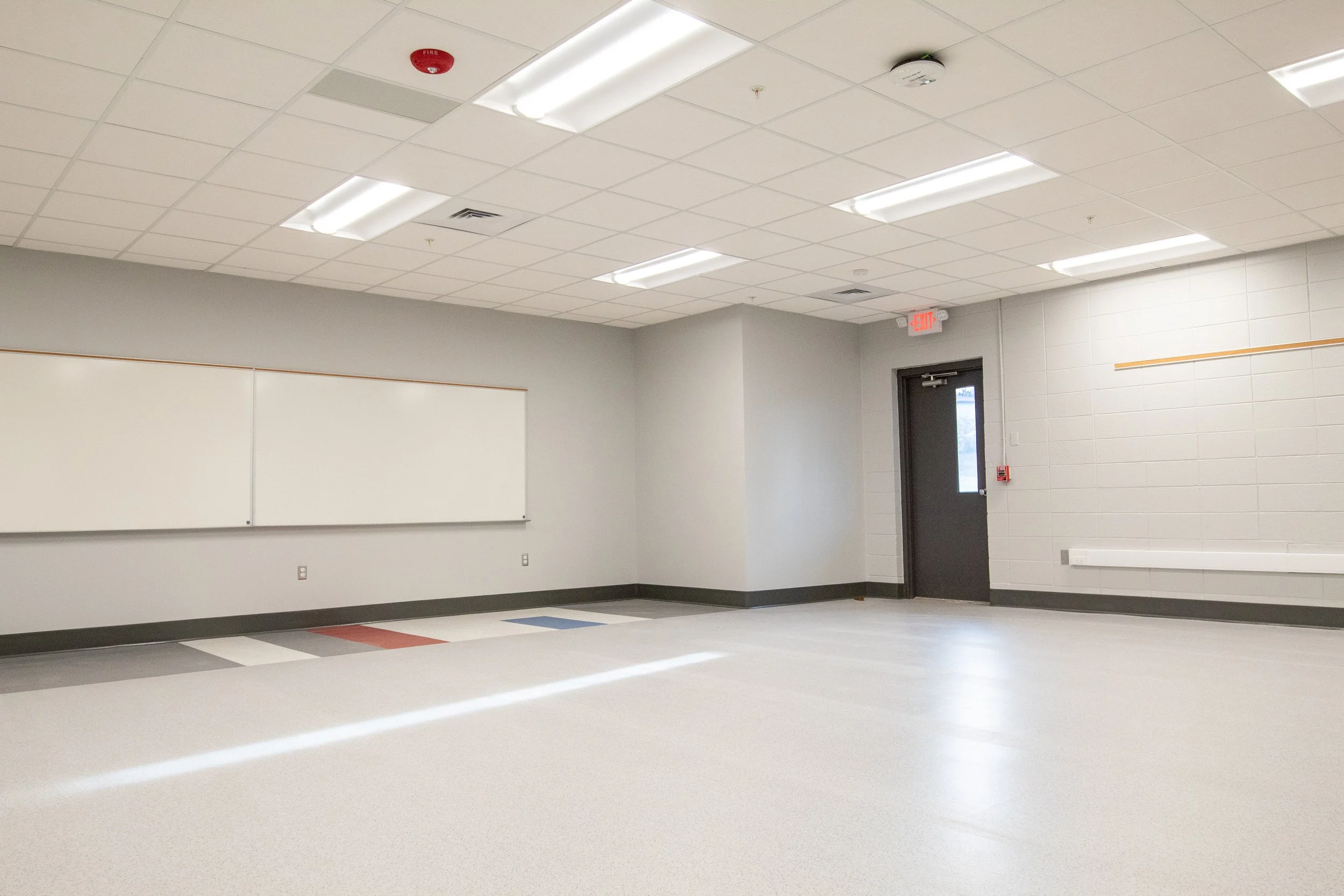 Empty classroom with white walls, ceiling tiles, fluorescent lights, a whiteboard, a closed exit door, and a fire alarm on the ceiling.