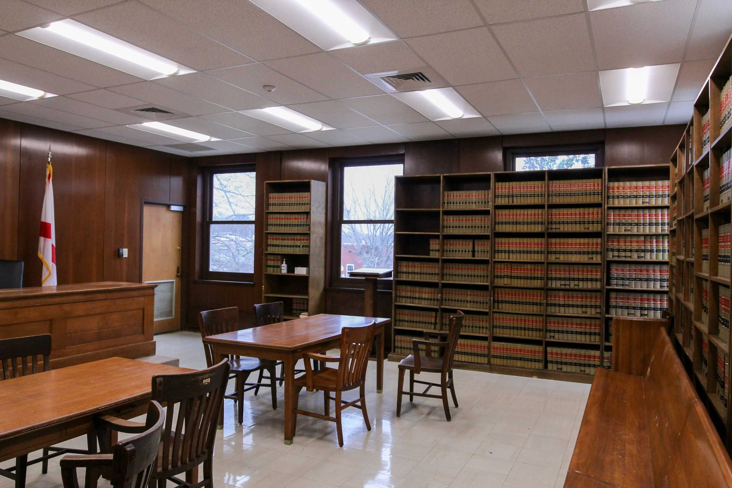 A room with wooden walls and multiple bookshelves filled with law books, wooden tables and chairs, windows with view of trees outside, and an American flag in the corner.