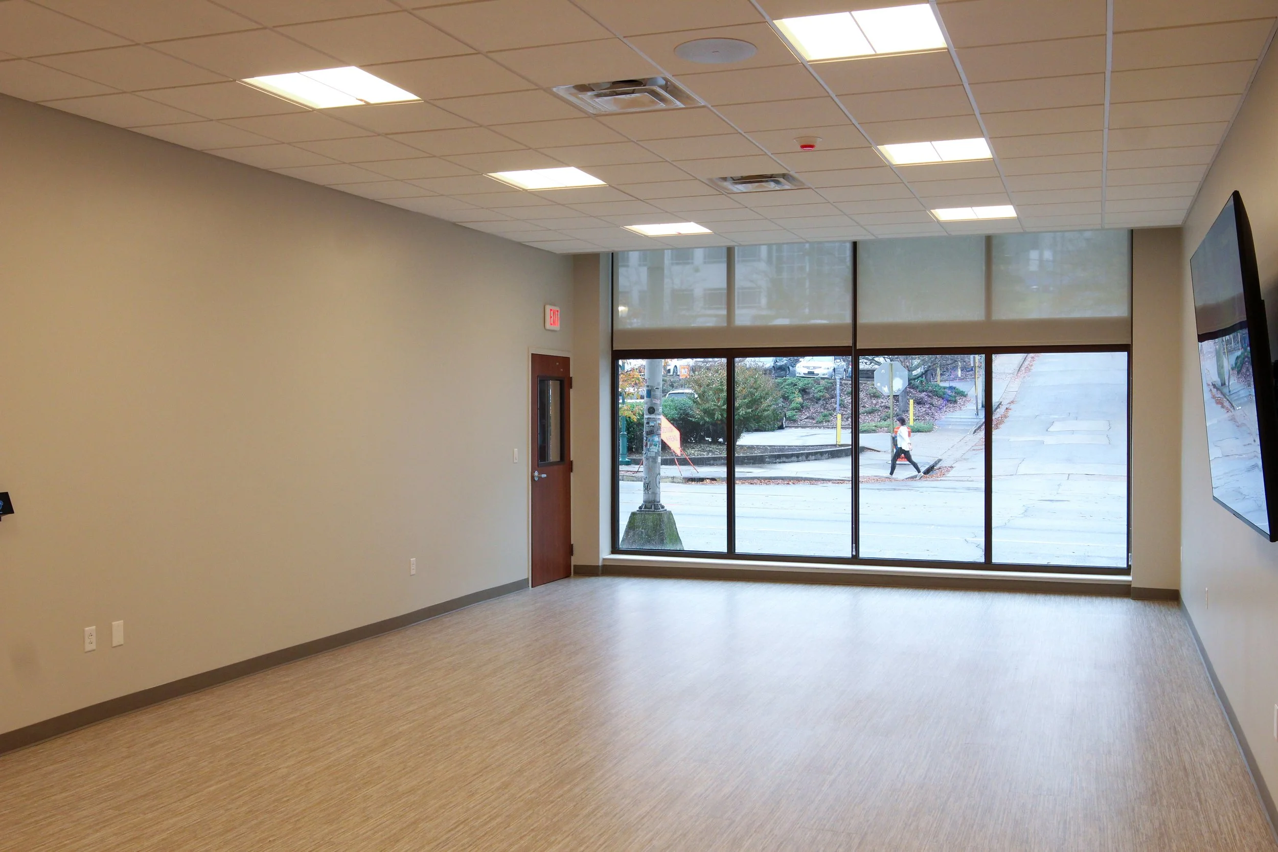 Empty office room with beige carpet, white walls, large window with a street view, ceiling with fluorescent lights, door with a window, and a wall-mounted television.