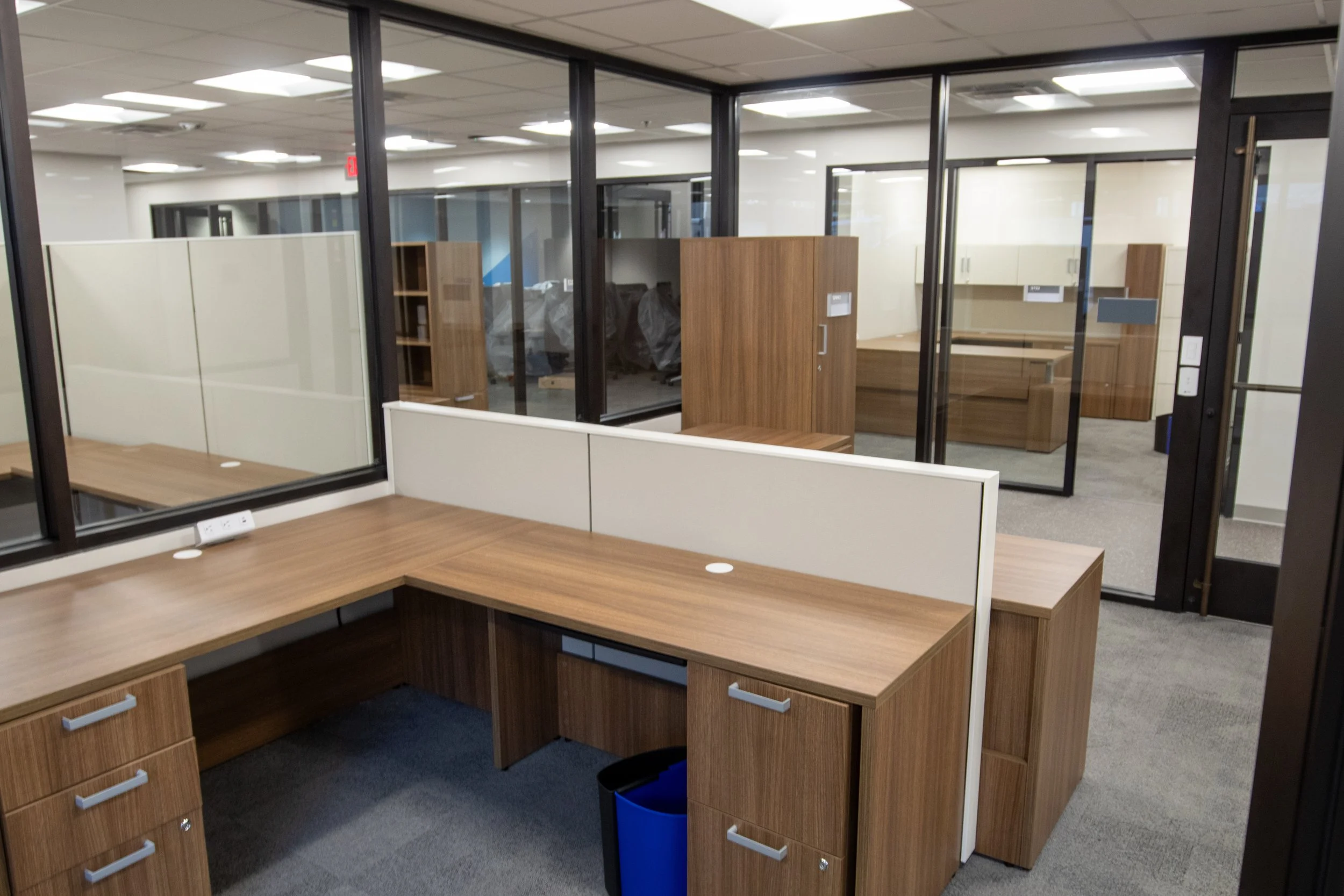 Empty office cubicles and desks with glass walls and wooden furniture in a modern workspace.