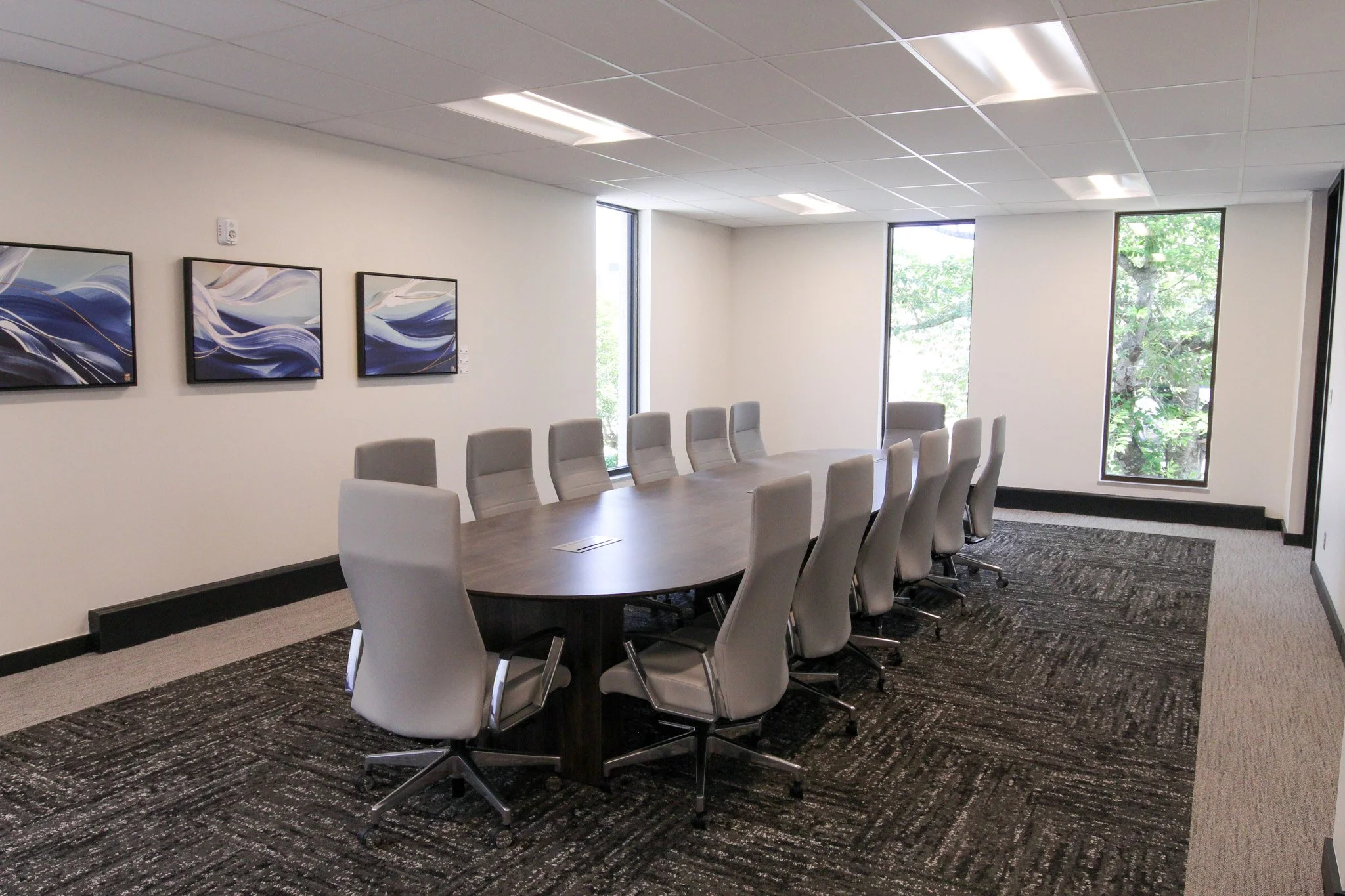 A modern conference room of Simmons Bank in Athens with a large oval table surrounded by beige office chairs. The room has three pieces of abstract artwork on the wall, large windows letting in natural light, white walls, and a textured dark carpet.