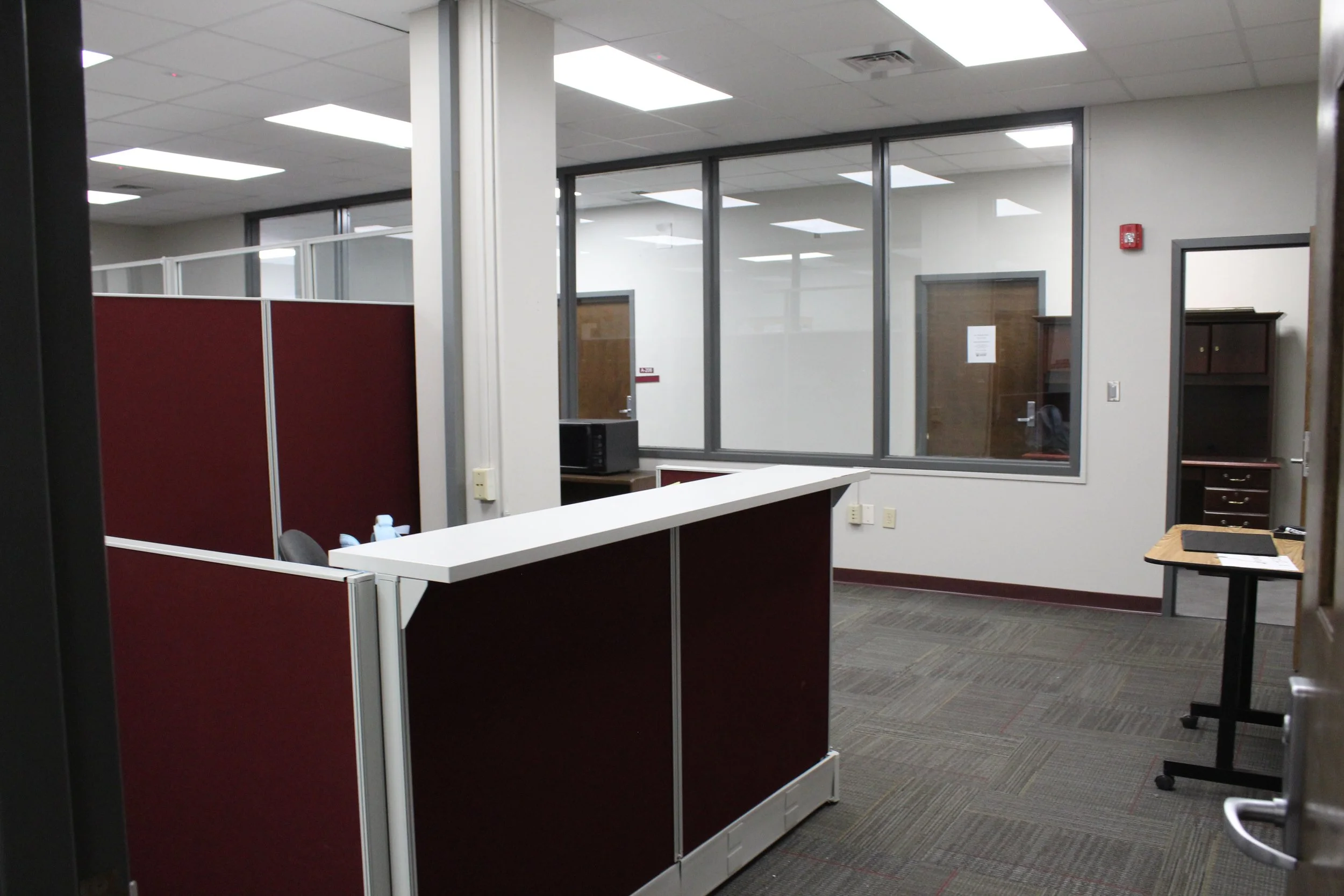 Empty office space with cubicles, a desk with a black mat, and glass-walled rooms.