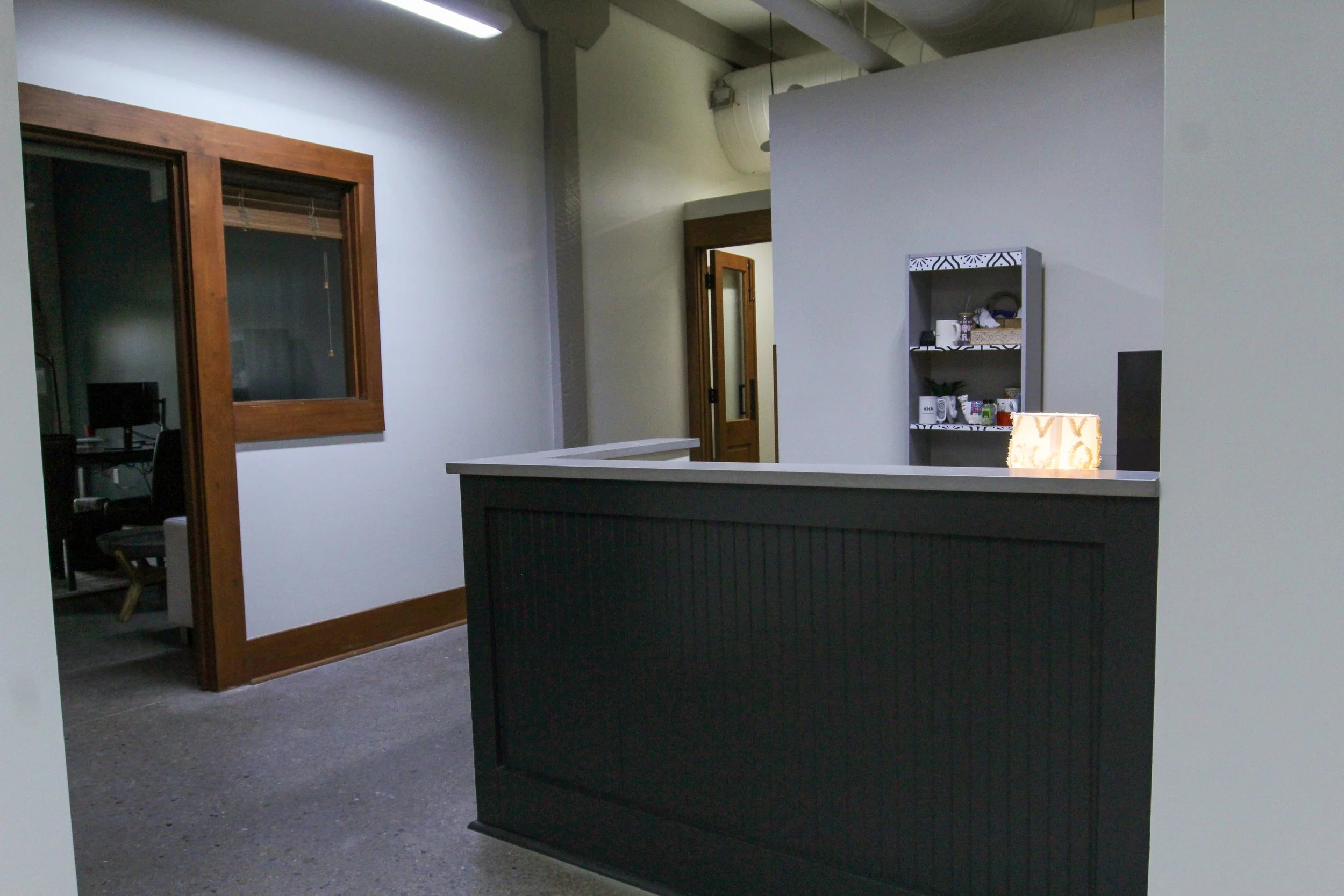 Office reception area with a black counter, a small shelf holding cups and other items, a door in the background, and a window in the wall on the left.