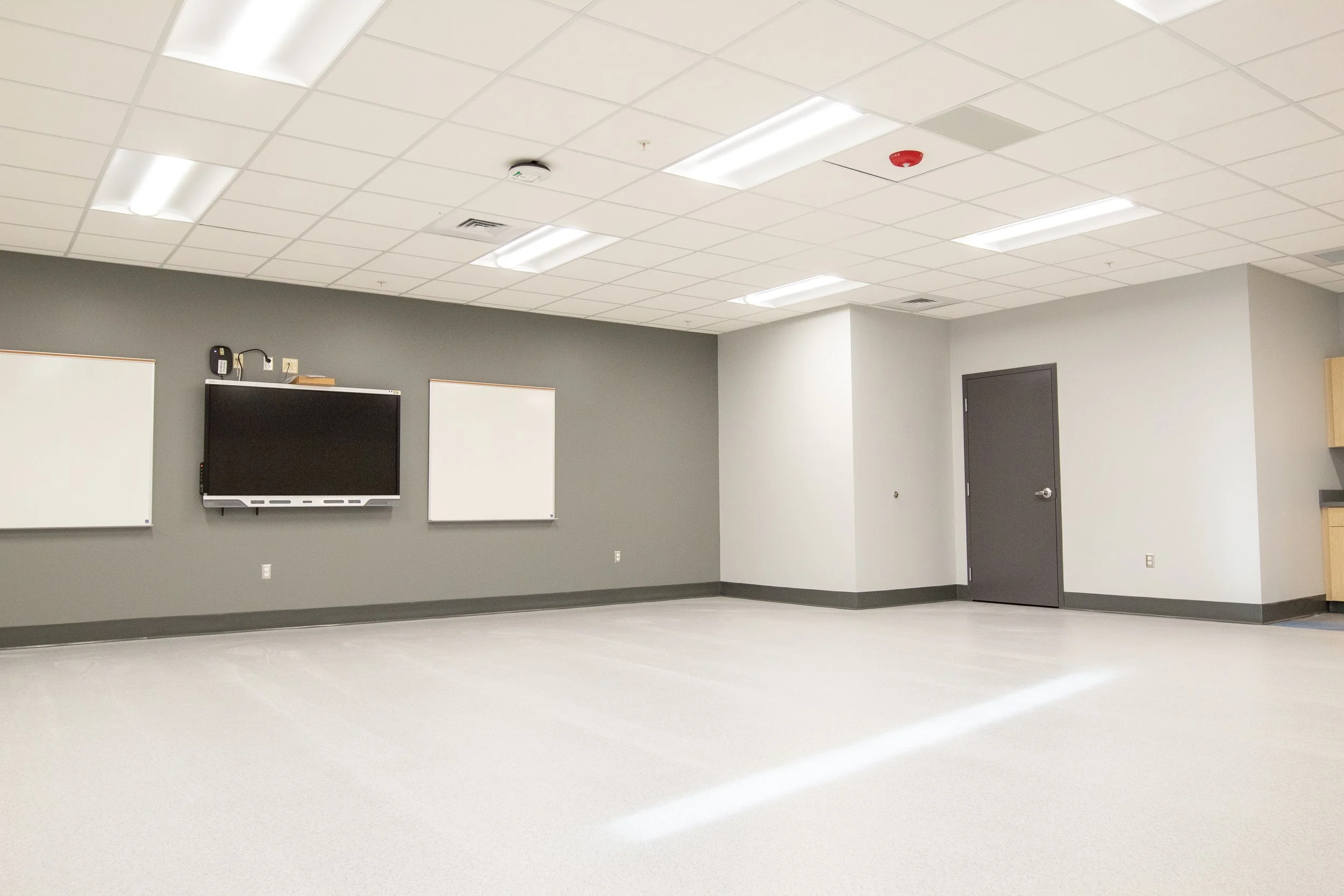 Empty classroom with gray walls, white ceiling, a mounted TV, two whiteboards, a gray door, and some cabinets in the corner.