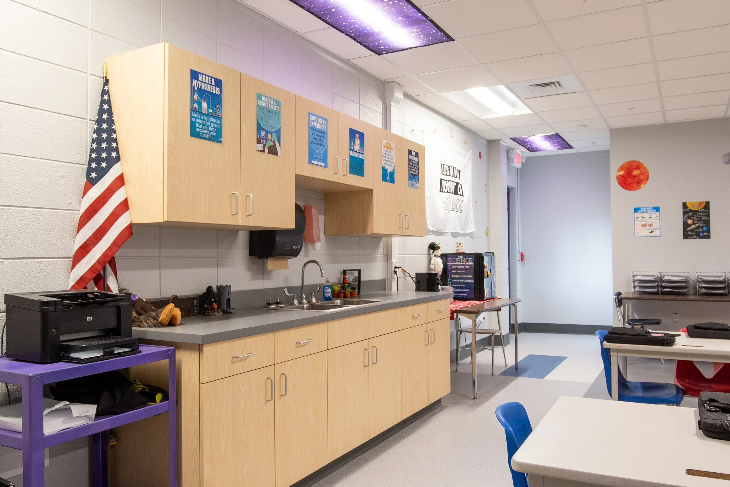 Classroom science area with cabinets, a sink, educational posters, American flag, and student desks.