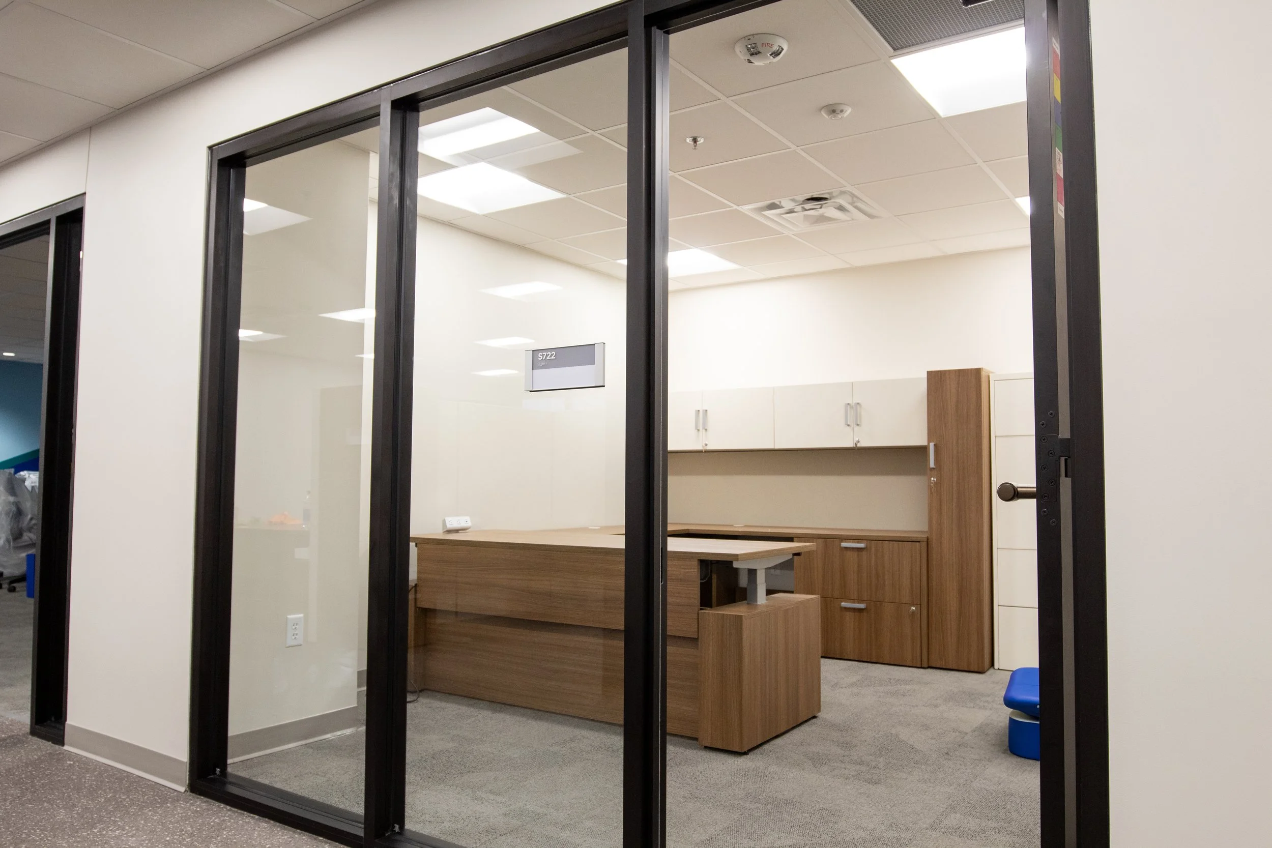 Empty office cubicle with wooden desk, white cabinets, beige walls, glass door, and ceiling lights.