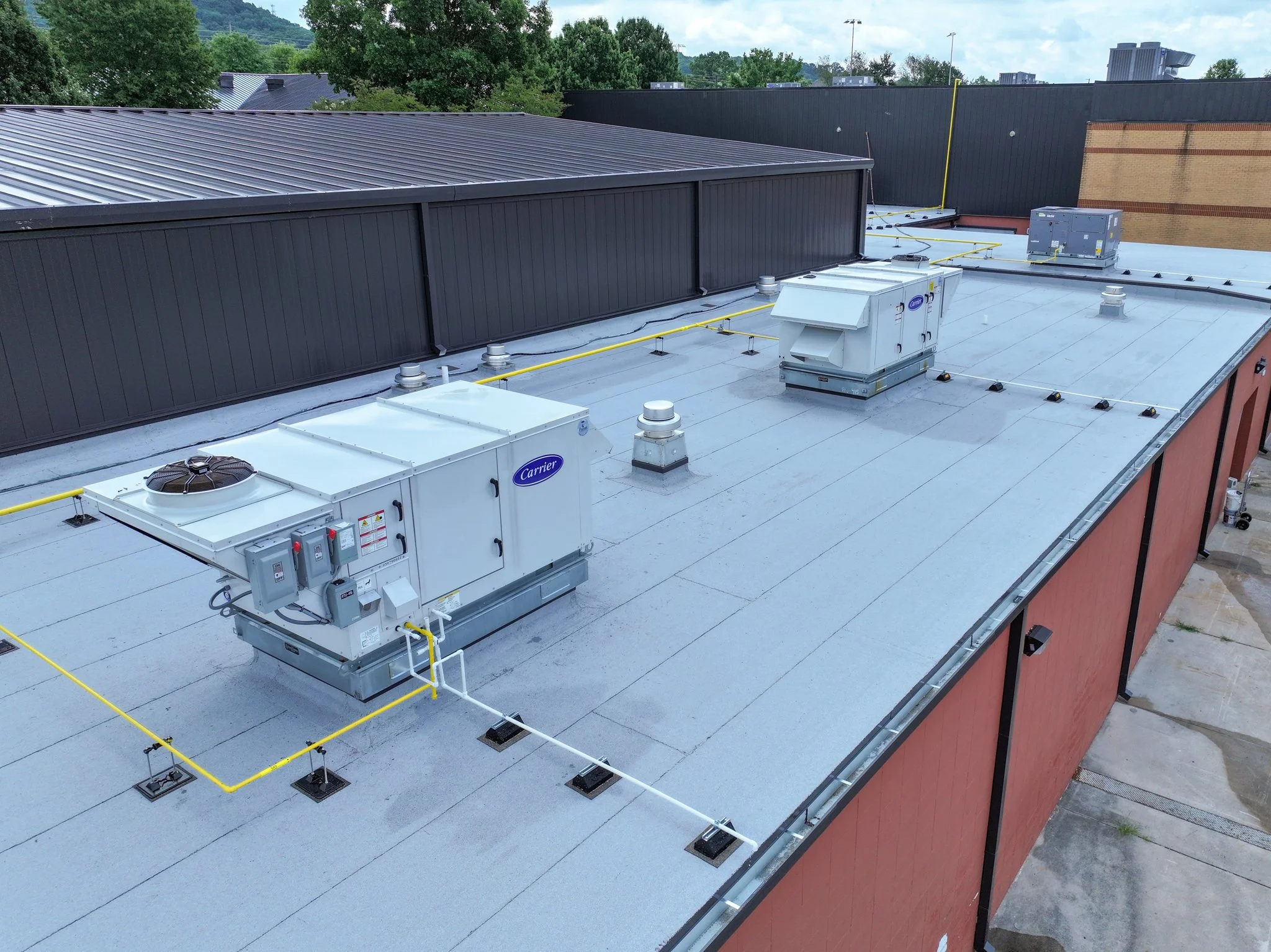 Rooftop view showing HVAC units, ventilation pipes, and black and gray metal siding with some trees and a cloudy sky in the background.