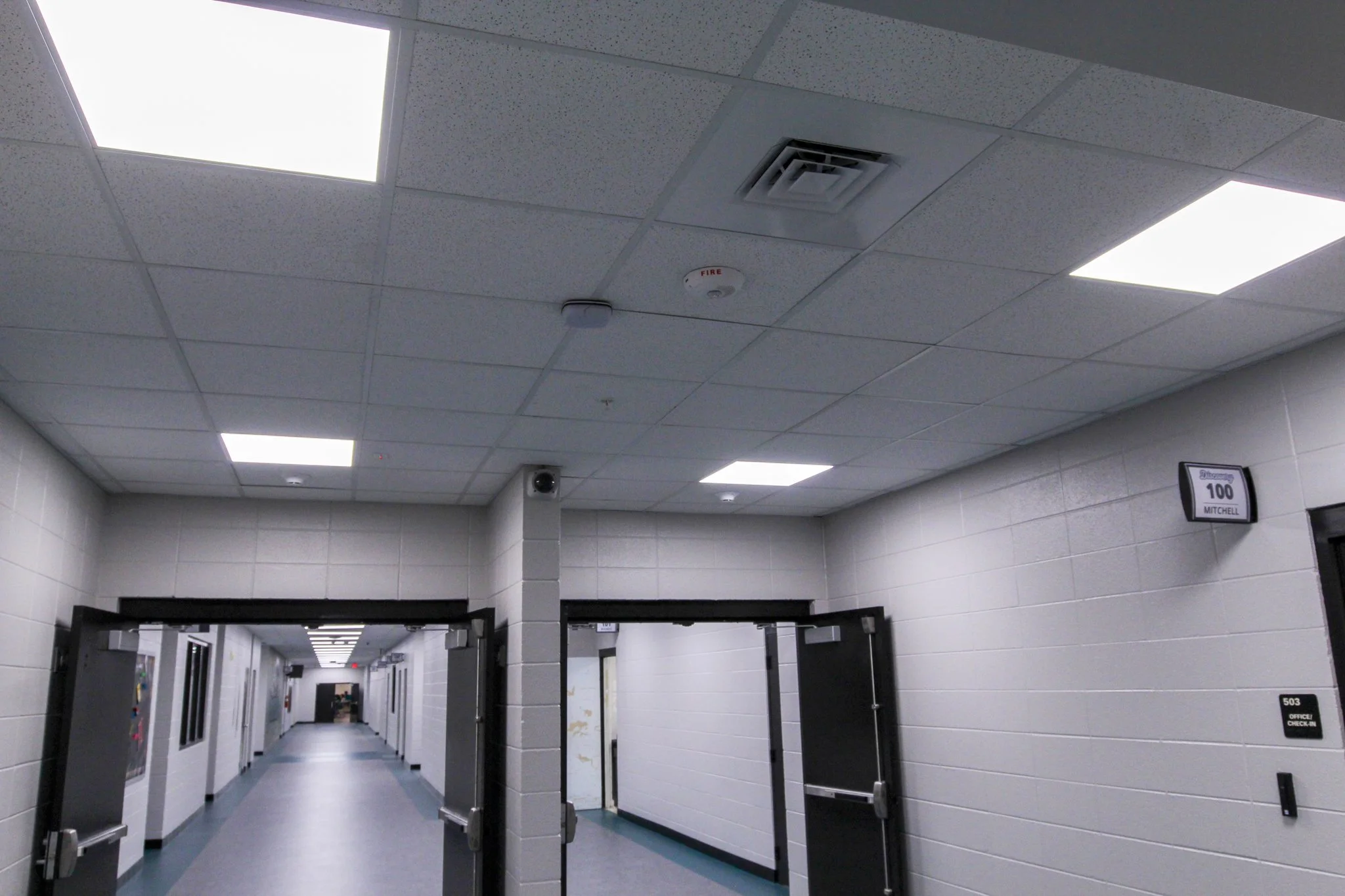 Hospital corridor with ceiling lights, fire alarm, ventilation system, and open doorways.