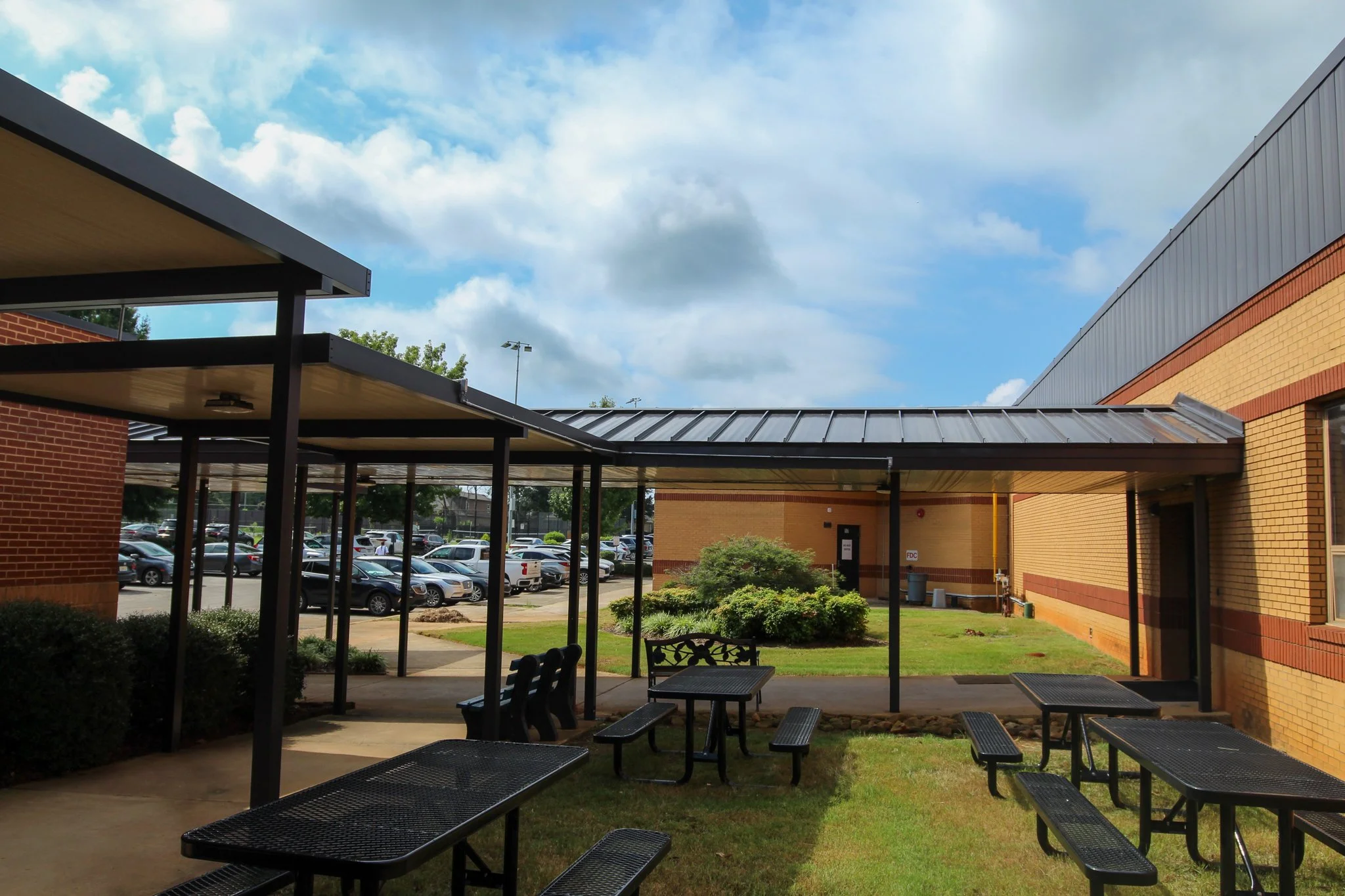 A view of a school or educational building with brick walls, metal roofing, and outdoor benches under a covered walkway, with a parking lot and blue sky with clouds in the background.