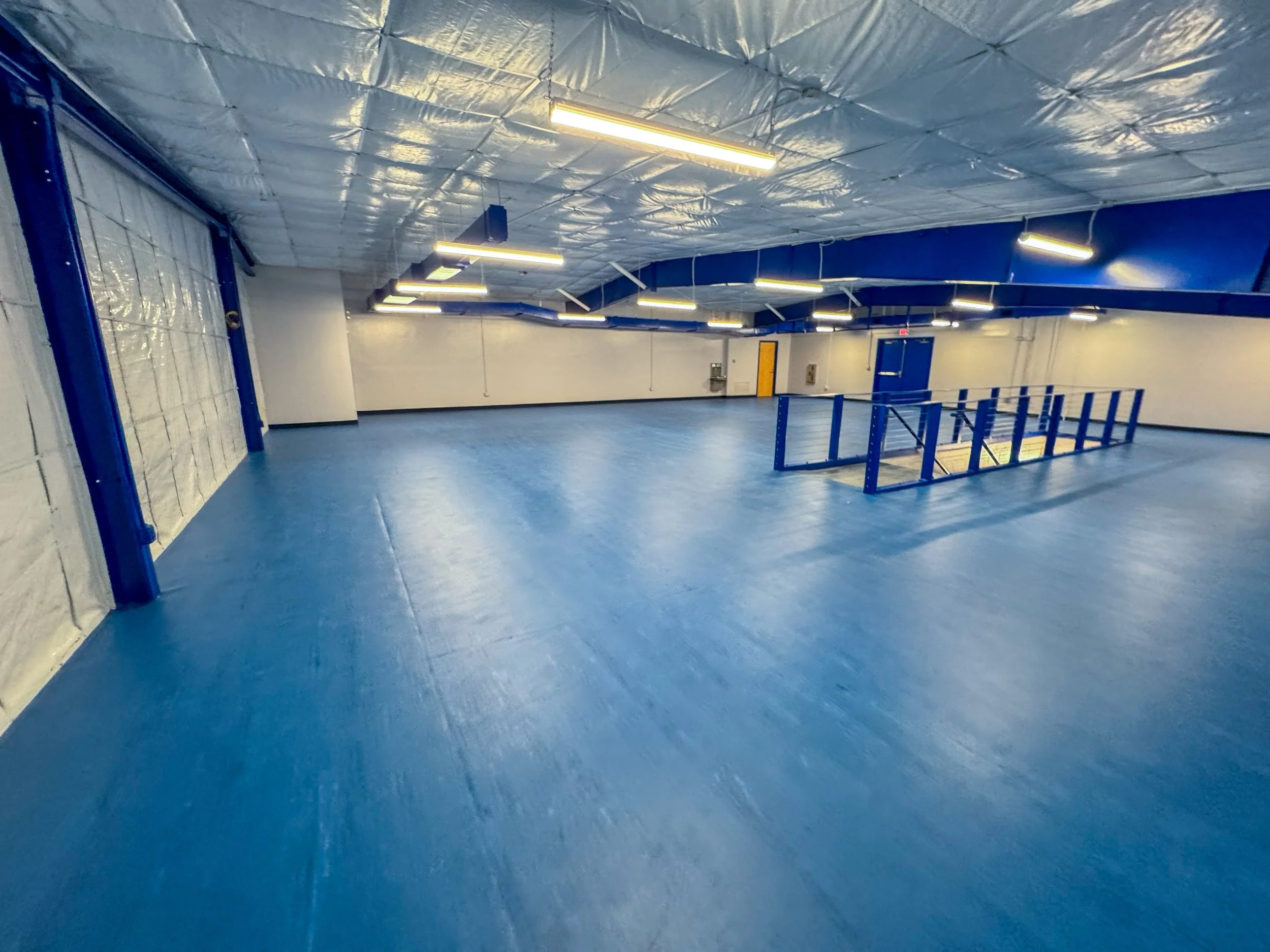 Empty indoor space with blue flooring, white and blue walls, blue railing around a staircase, ceiling with exposed insulation, and fluorescent lighting.